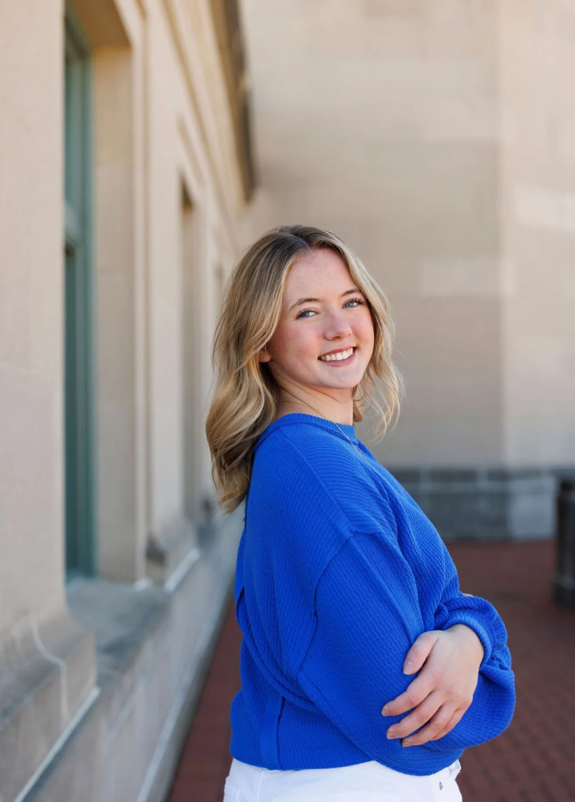 A young woman with blond hair, smiling, wearing a blue sweater, standing outdoors near a beige building with windows, with arms crossed.
