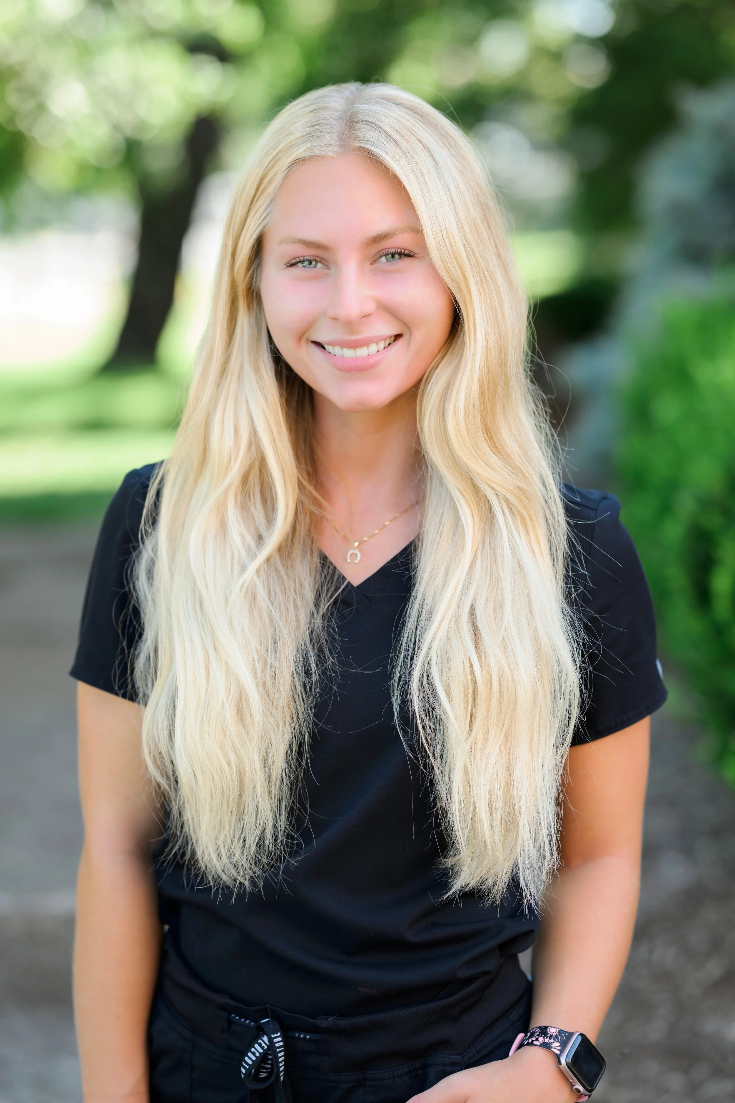 A young woman with long blonde hair, wearing a black shirt and a smartwatch, standing outdoors with a background of green trees and blurred greenery.
