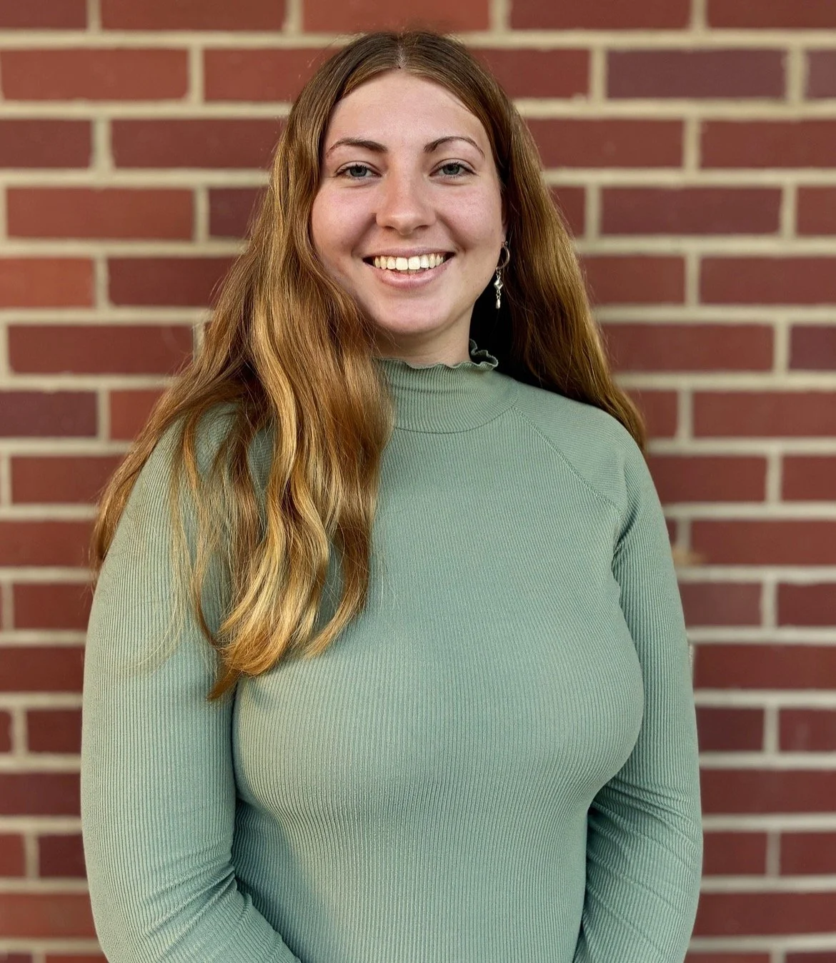 A young woman with long red hair, smiling, standing in front of a red brick wall, wearing a green ribbed long-sleeve shirt.