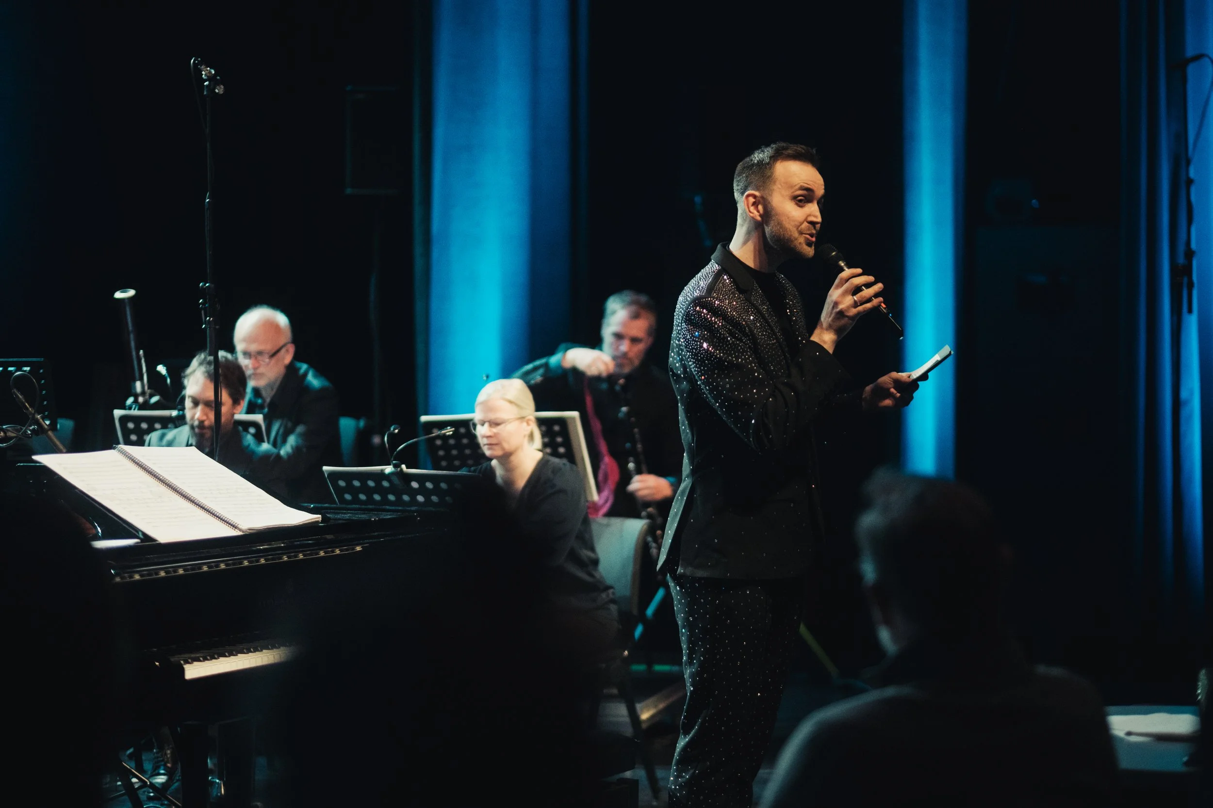 En mann i svart dress som står på en fjelltopp med utsikt over en innsjø og grønne åser i bakgrunnen.