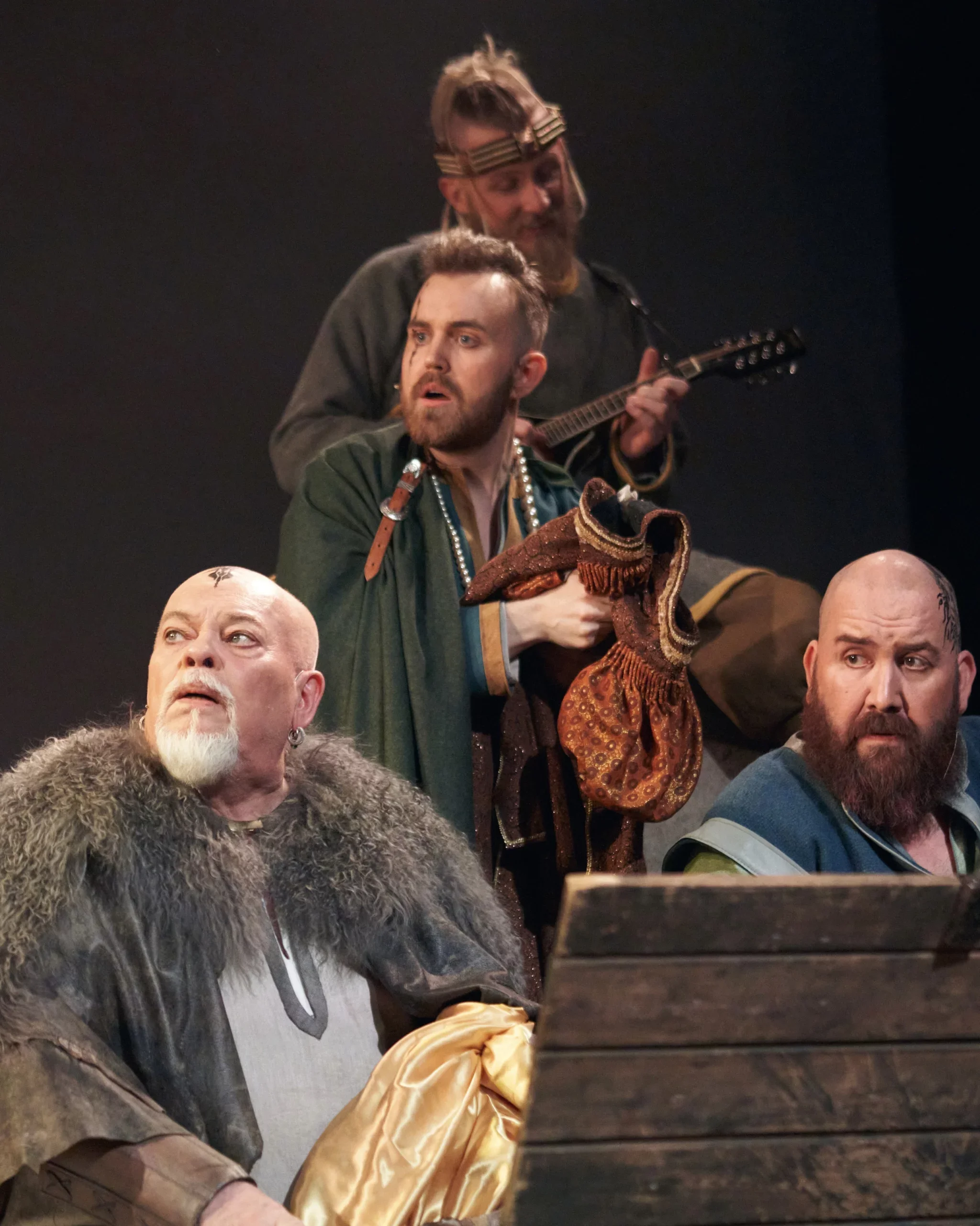 Fire menn iført kostymer, med to av dem med skjegg, en med hår og en med en stor pelsjakke, ser på noe utenfor bildet, mens en annen mann spiller en gitar i bakgrunnen.