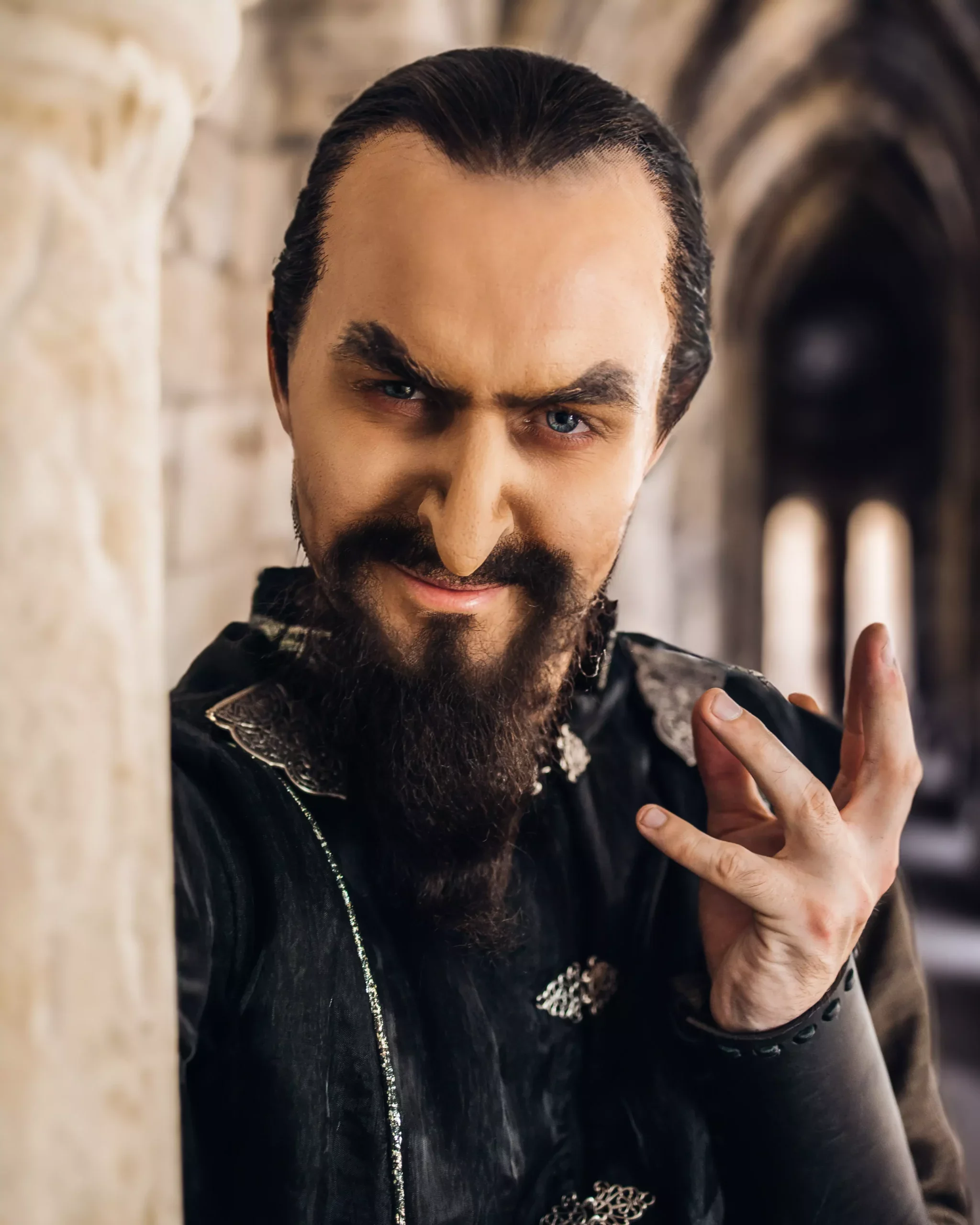 En mann med mørkt hår og fullt skjegg kler svart lærjakke med sølvdetaljer, står i en arkeologisk bygning med buede vicer. Han har et lekent uttrykk og gjør en håndbevegelse.
