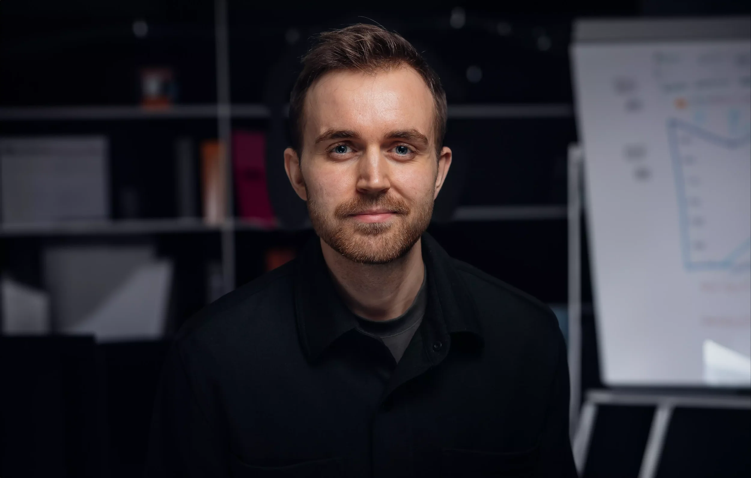 Portrett av en mann med mørkt hår og skjegg, iført svart skjorte, foran en presentasjonsvegg med diagram.