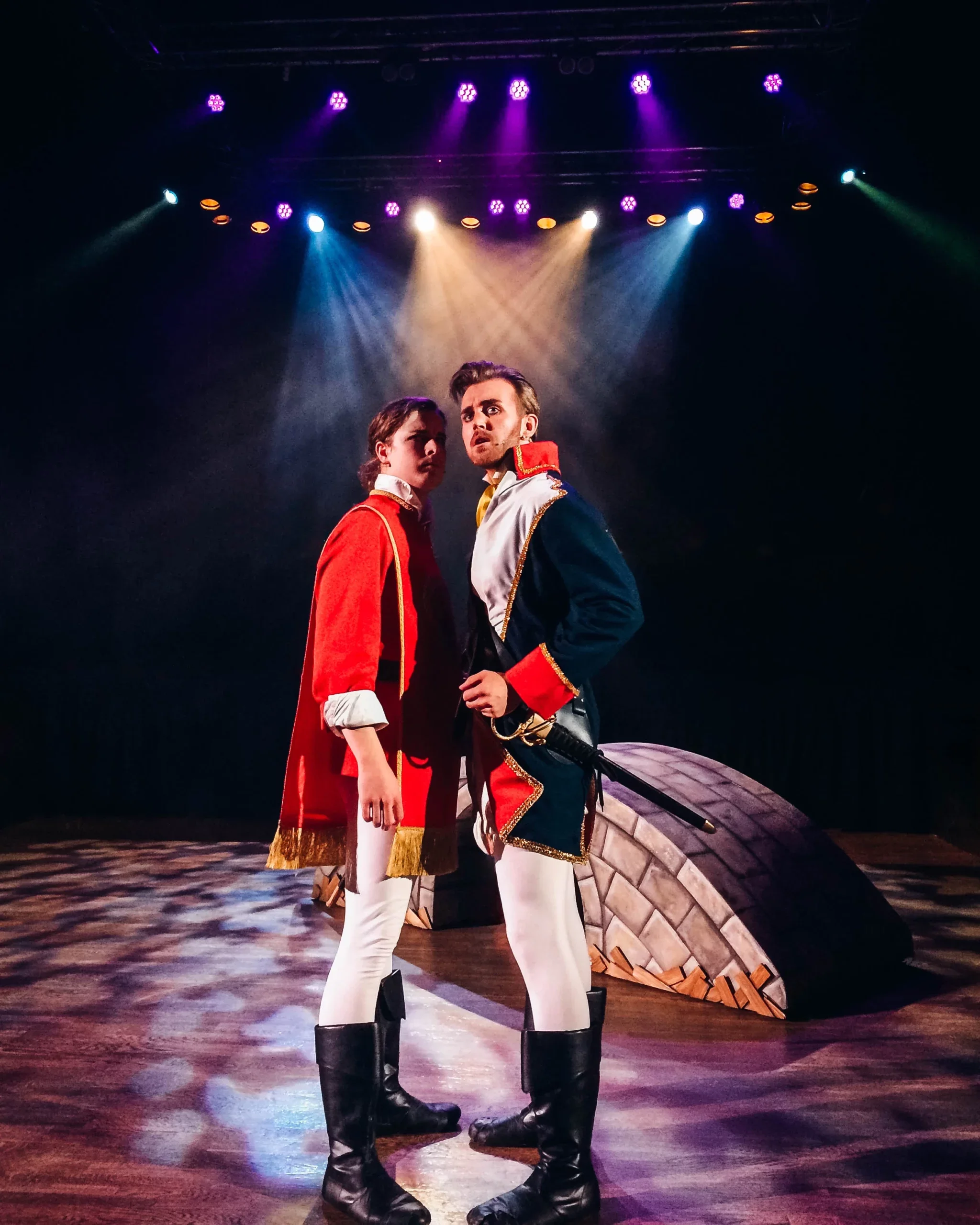 To skuespillere i historiske militære kostymer på scenen under en forestilling, med teaterlys over scenen.