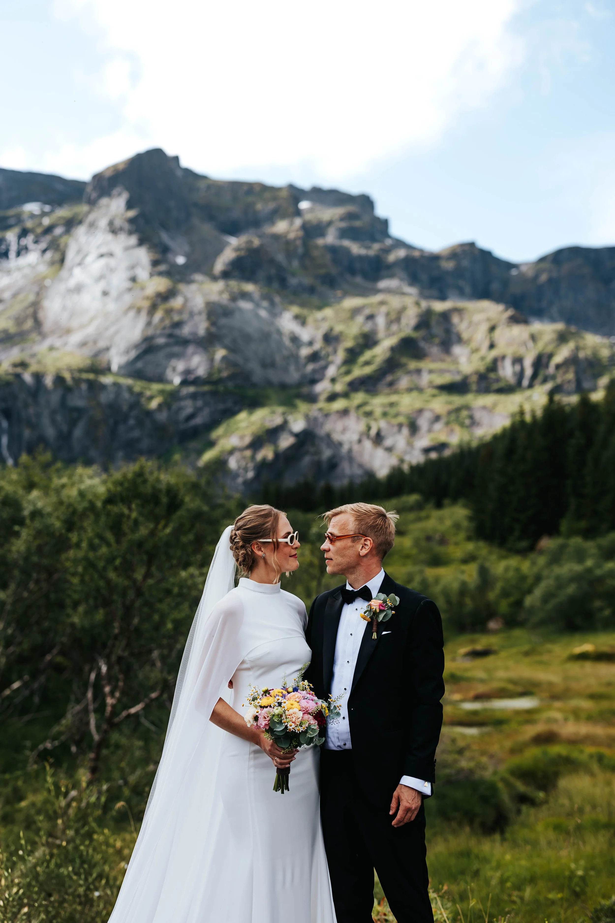 En brud og brudgom står ansikt til ansikt i en naturskjønn utendørs setting med fjell og skog i bakgrunnen.