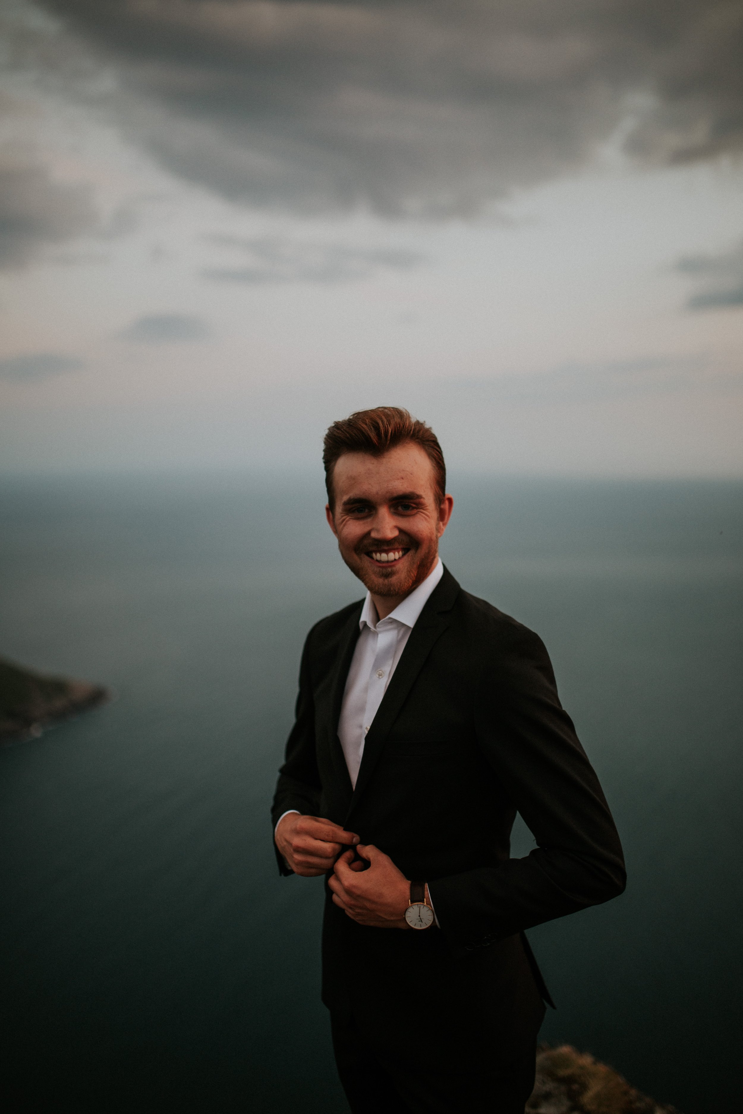 Smilende mann i svart dress med hvit skjorte, stående på klippe med utsikt over vann og mørke skyer.