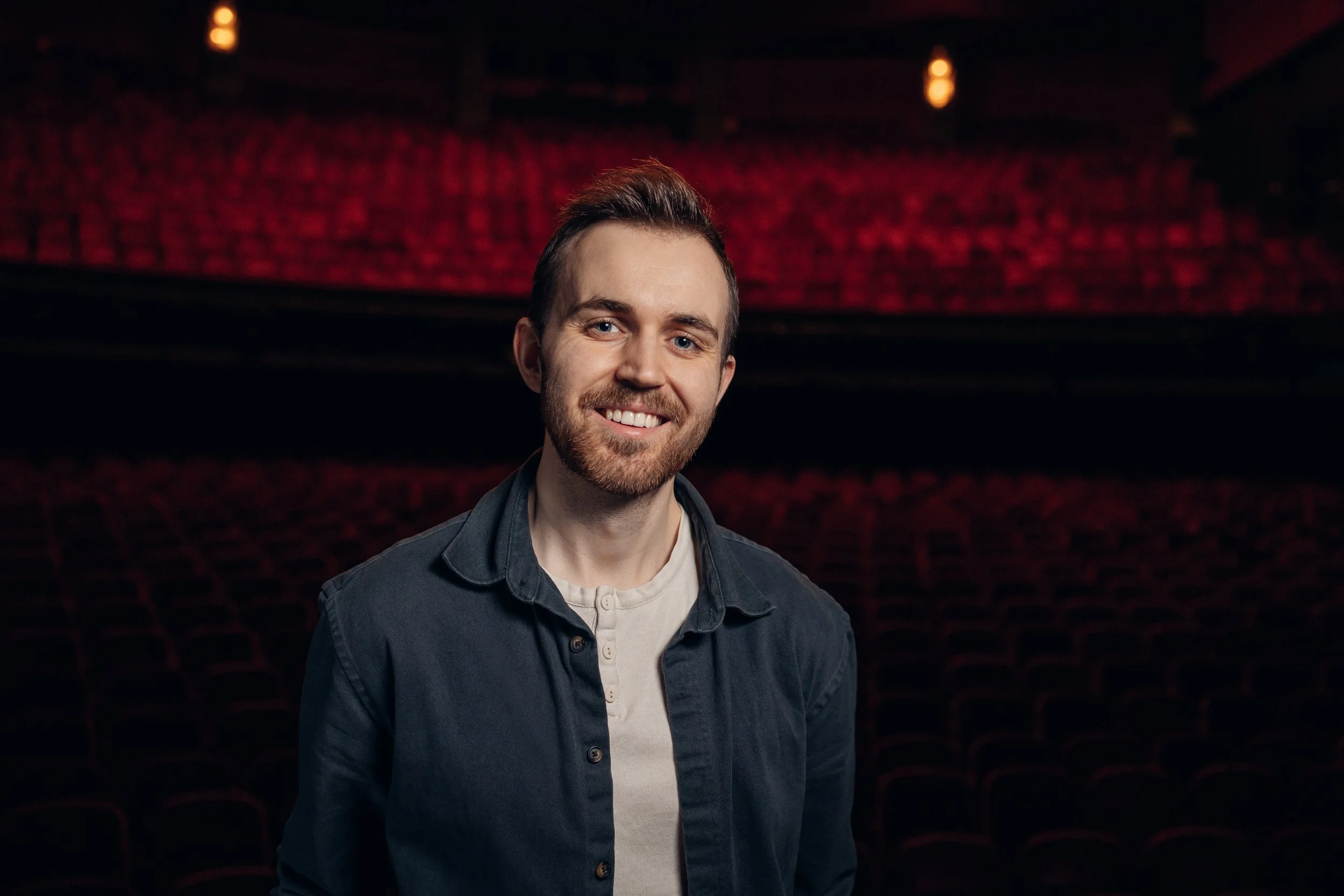 En ung mann med bart og lys hår står i et teater med røde stoler i bakgrunnen.