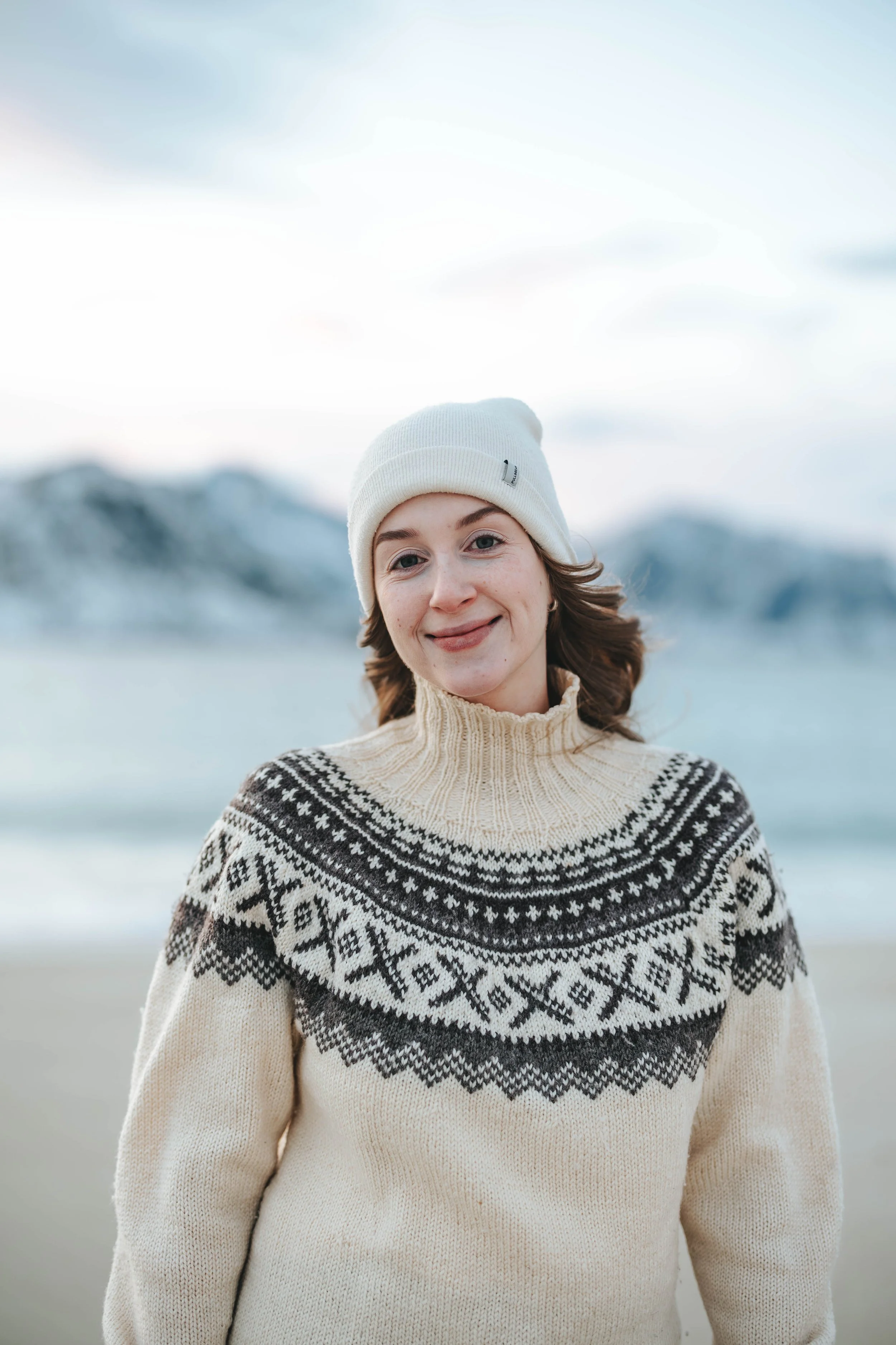 Et smilende kvinne med brunt, bølget hår, iført en hvit ullhatt og en beige og svart mønstret genser, står foran snødekte fjell og en vannløsning.