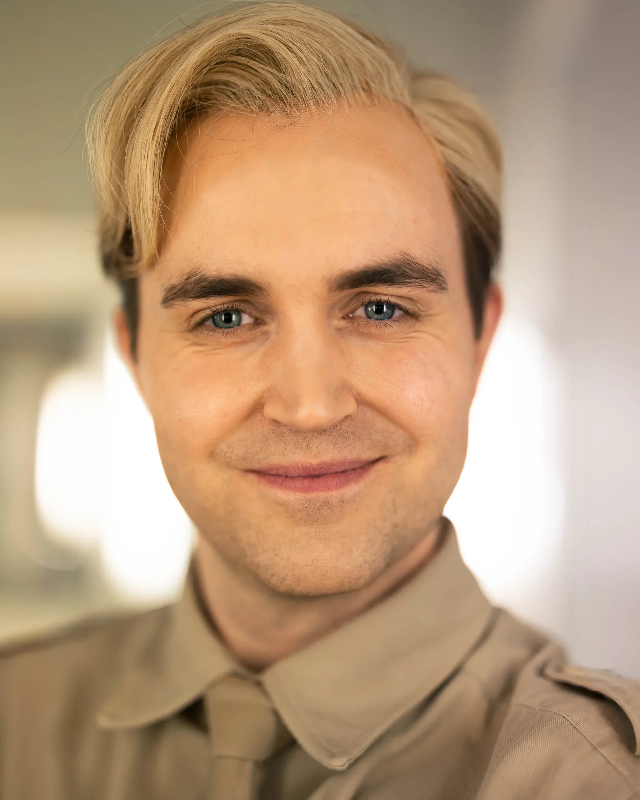 Et nærbilde av en mann med blond, sidekappet hår og blå øyne, smilende, iført en beige uniform med epauletter.