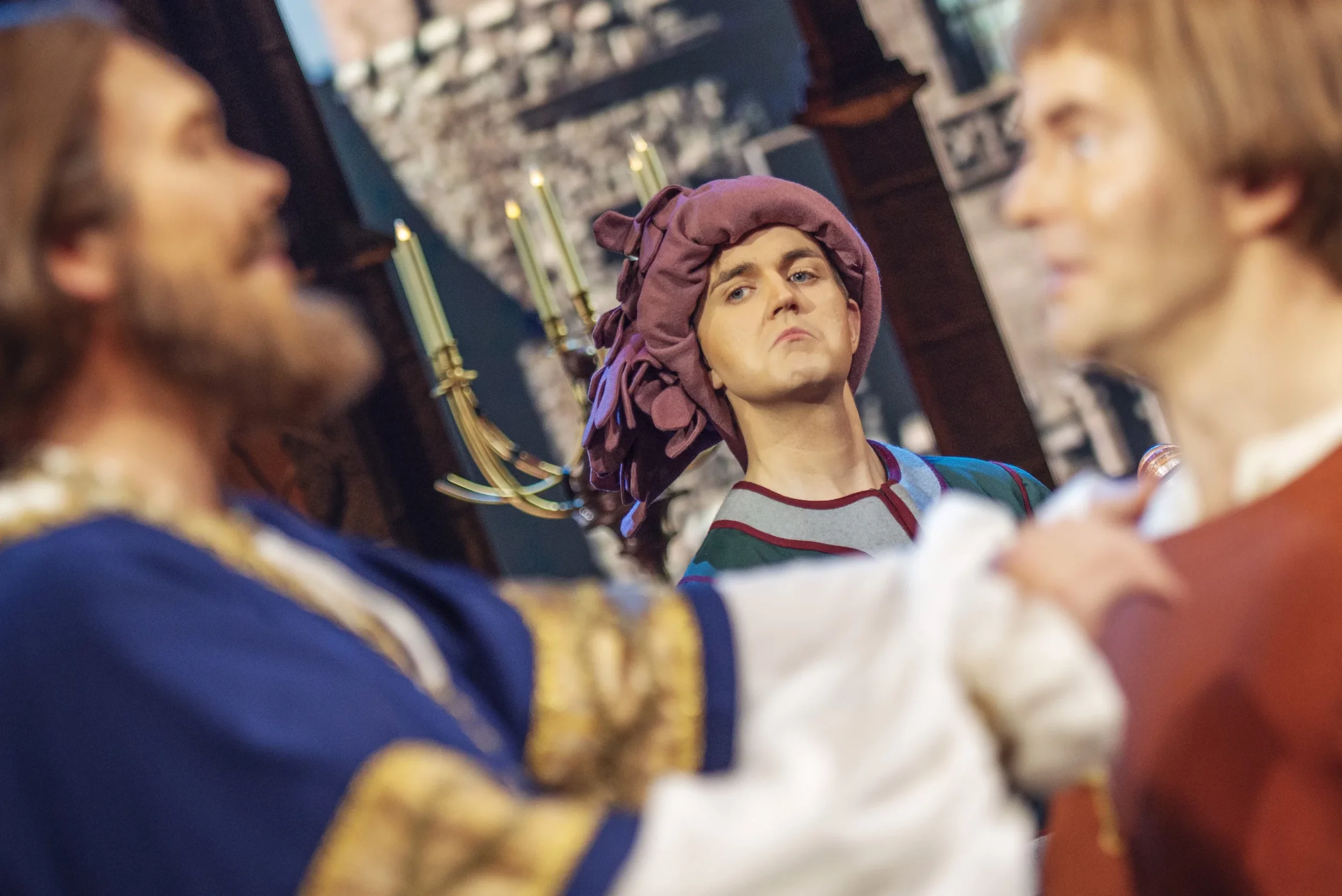 En scene med tre personer i historisk kostyme, med en person i midten som ser skeptisk eller misfornøyd ut.