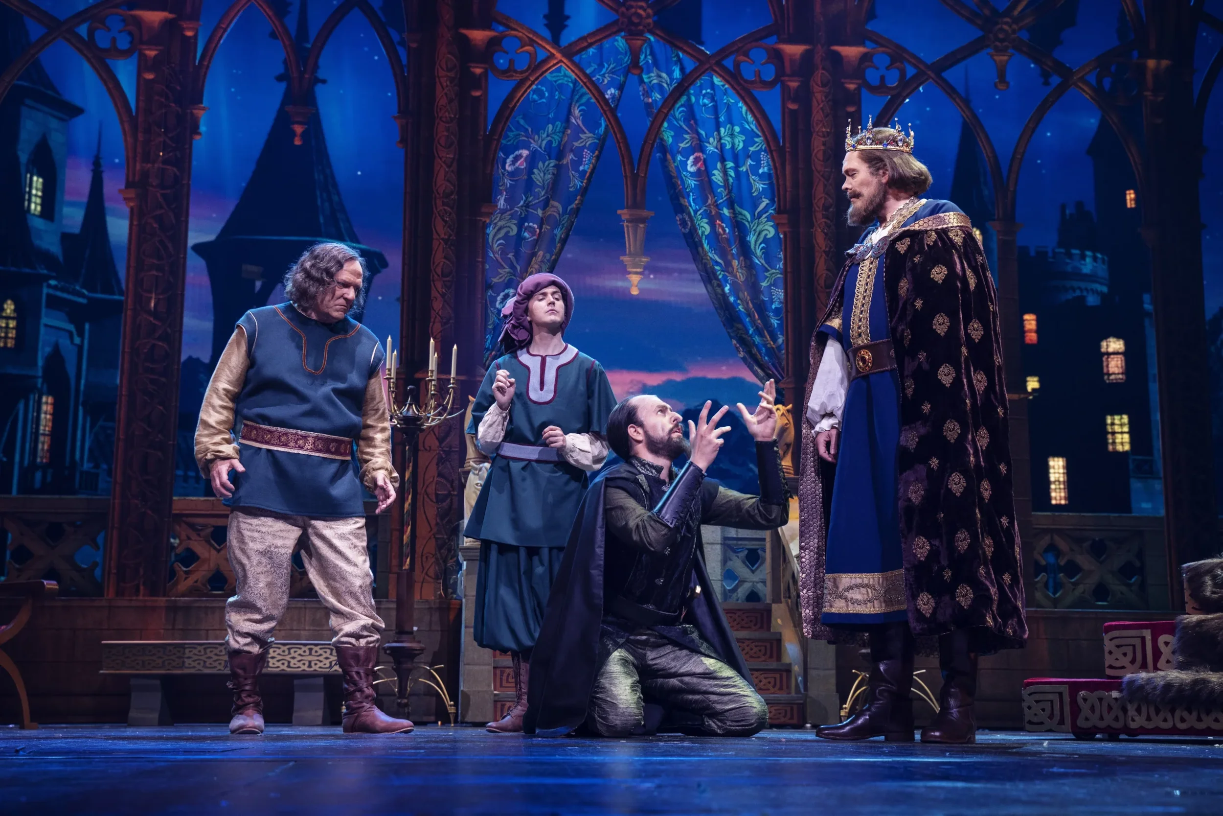 En scene fra en teaterforestilling med fem skuespillere i kostymer, en av dem er kongen med krone, som står foran en bakgrunn som ser ut som en middelalderslott.