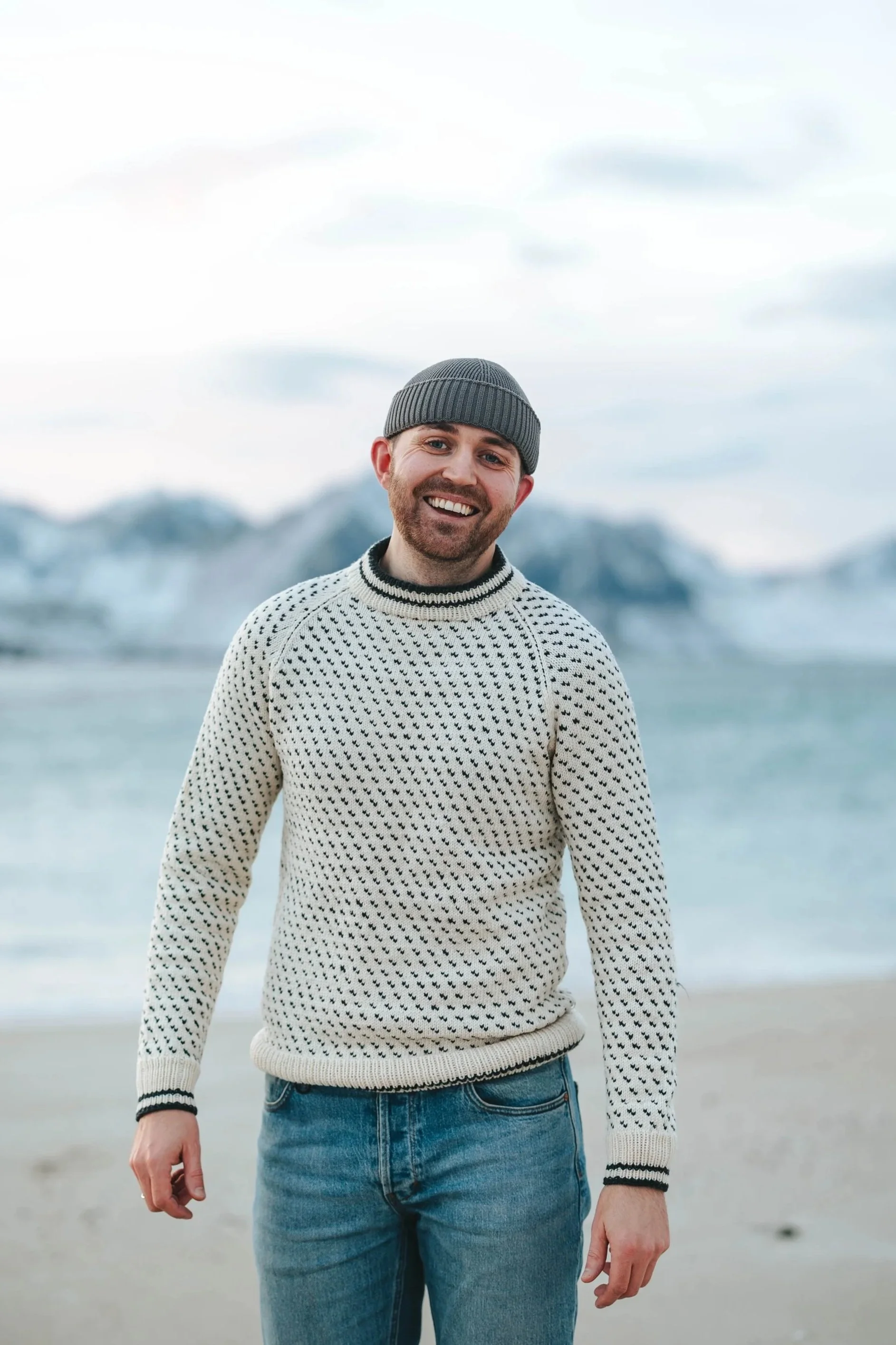 Smilende mann i stripete genser og beanie, står på en strand med fjell i bakgrunnen.
