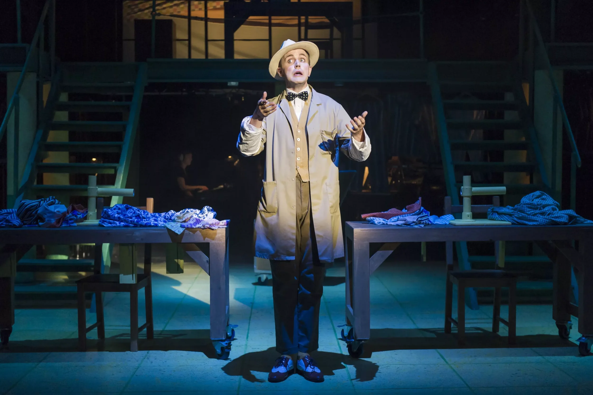En skuespiller står på en teaterscene iført en beige frakk, en hvit hatt, og en svart-hvitt sløyfe, og gestikulerer med hendene.