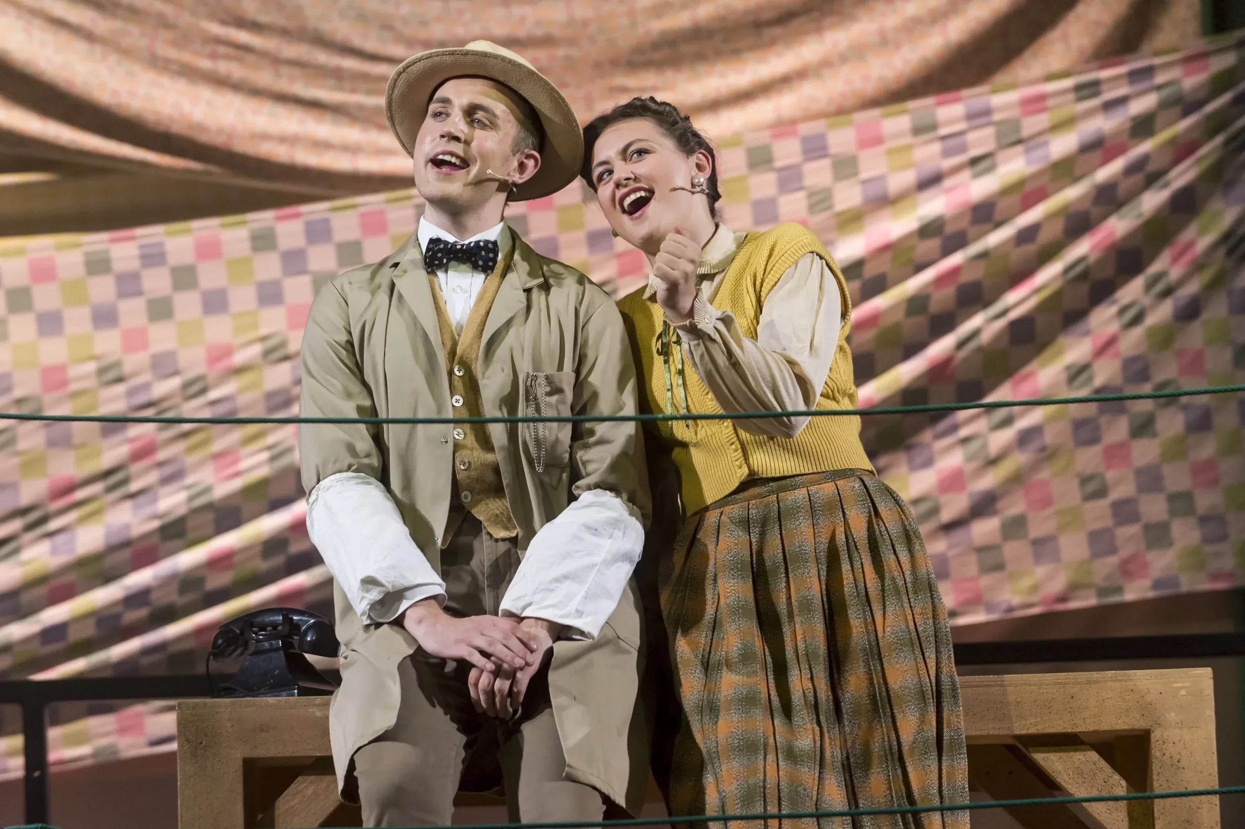 To personer på scenen, en mann og en kvinne, i kostymer, under en forestilling med fargerikt bakteppe. Mannen sitter på en kasse med en telefon foran seg, kvinnelig figur står ved siden av ham.