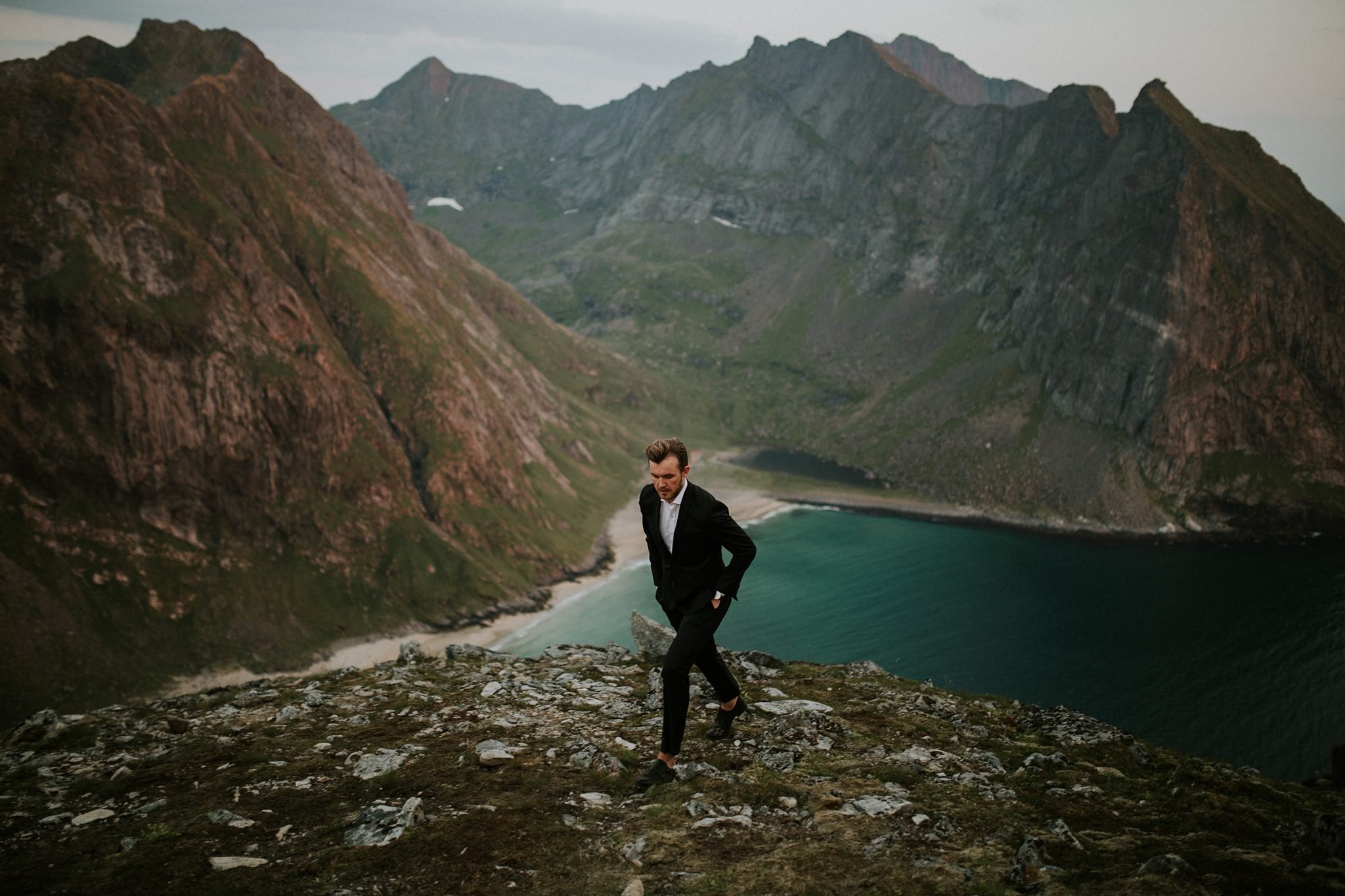 En mann i formelt antrekk som går opp en skråning med fjell i bakgrunnen og en innsjø under.