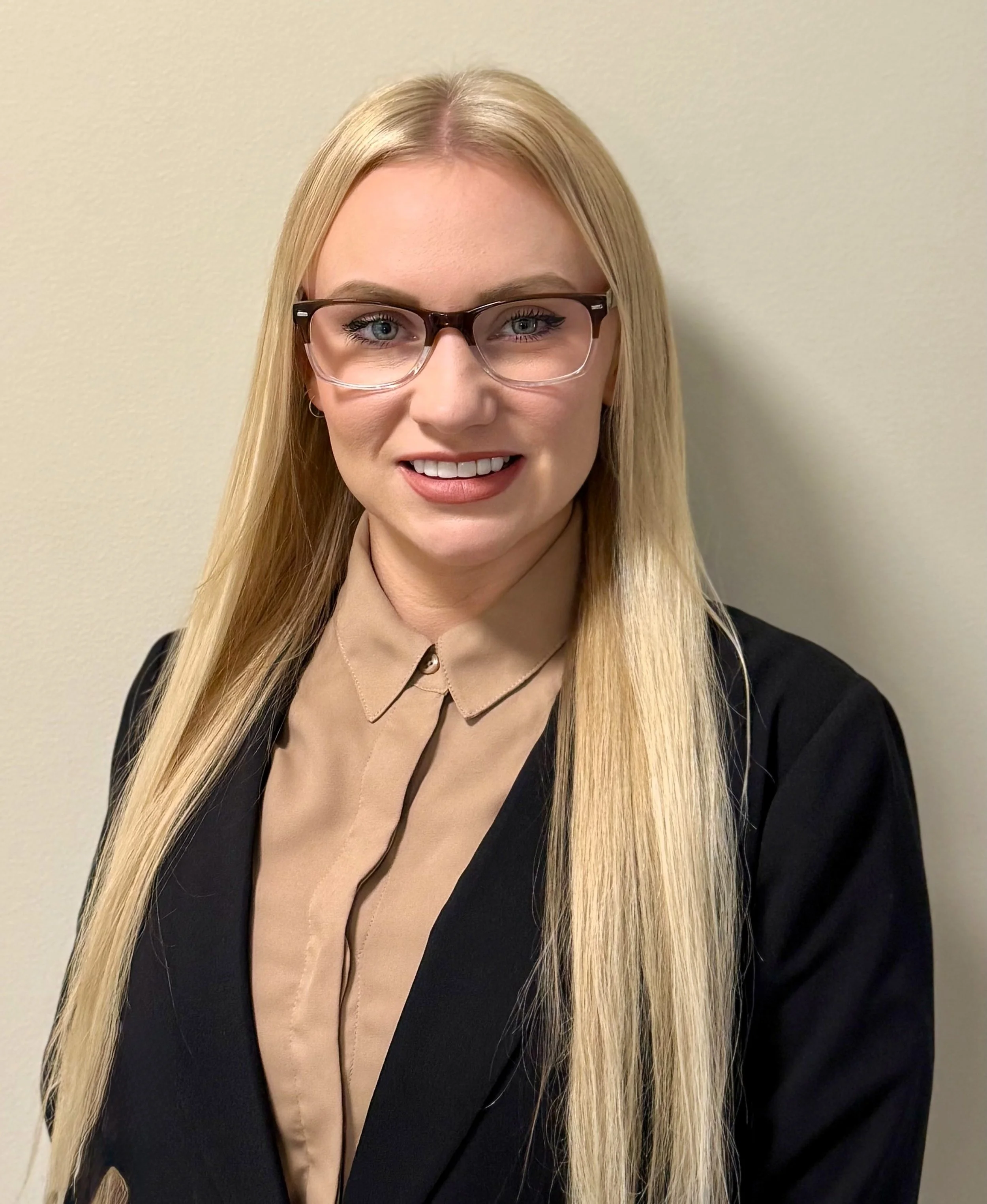 A woman with long blonde hair, wearing glasses, a black blazer, and a beige blouse, smiling in front of a plain beige wall.