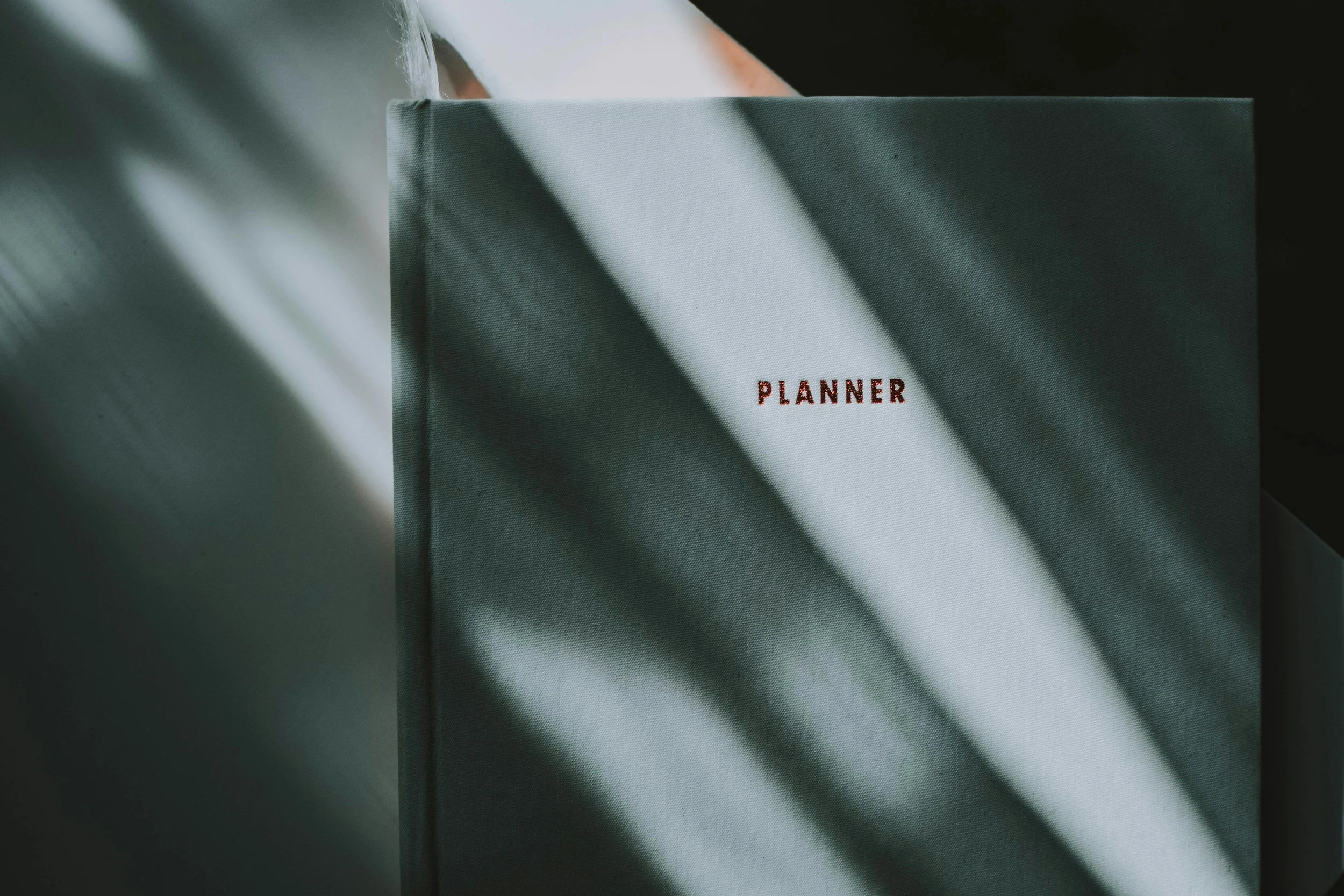 A green planner notebook with red embroidered text 'PLANNER' on the cover, placed on a surface with shadows and streaks of light.