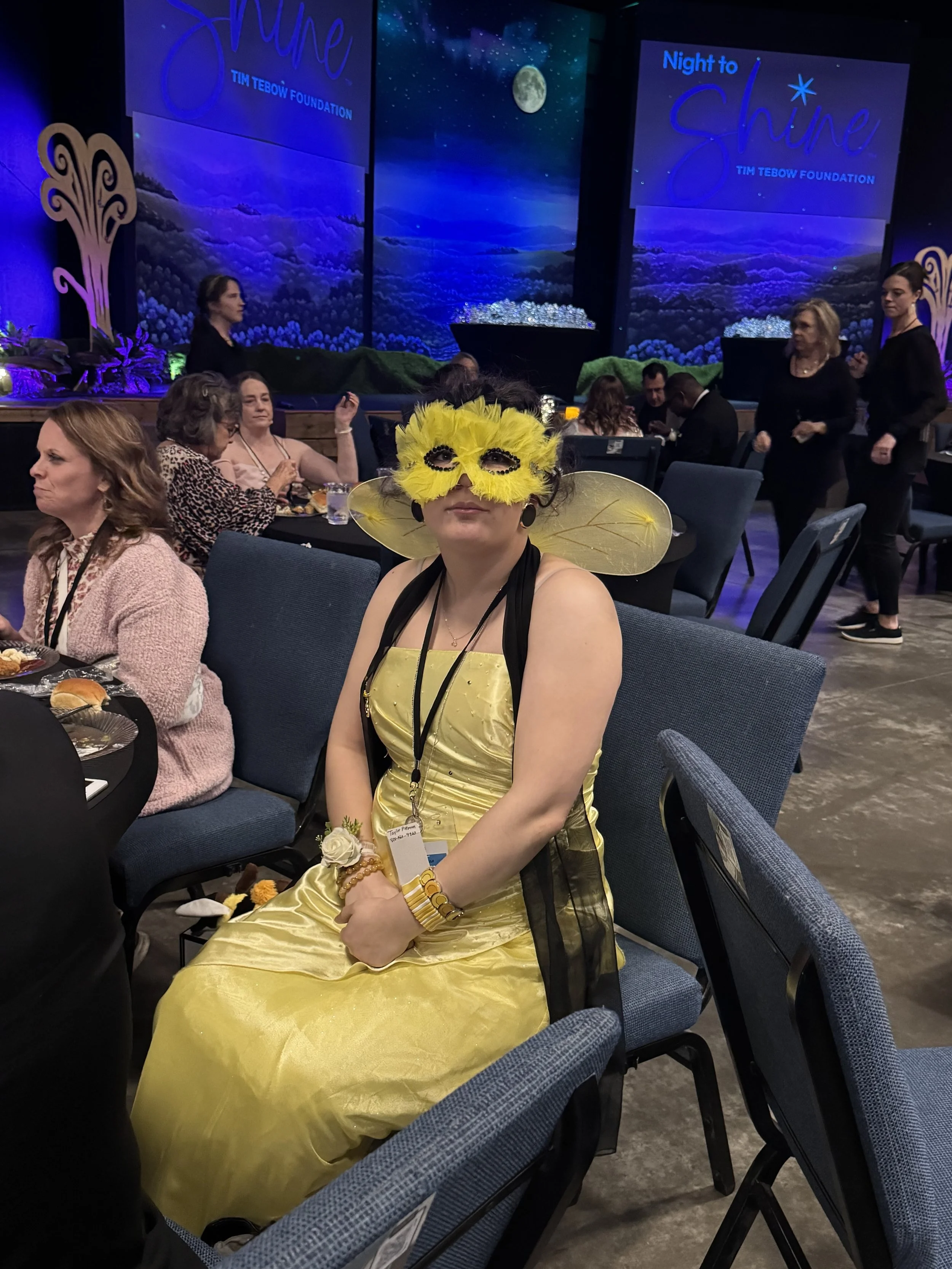 Woman wearing a yellow dress and yellow bird mask with wings, sitting at a banquet table during a formal event with a stage in the background.