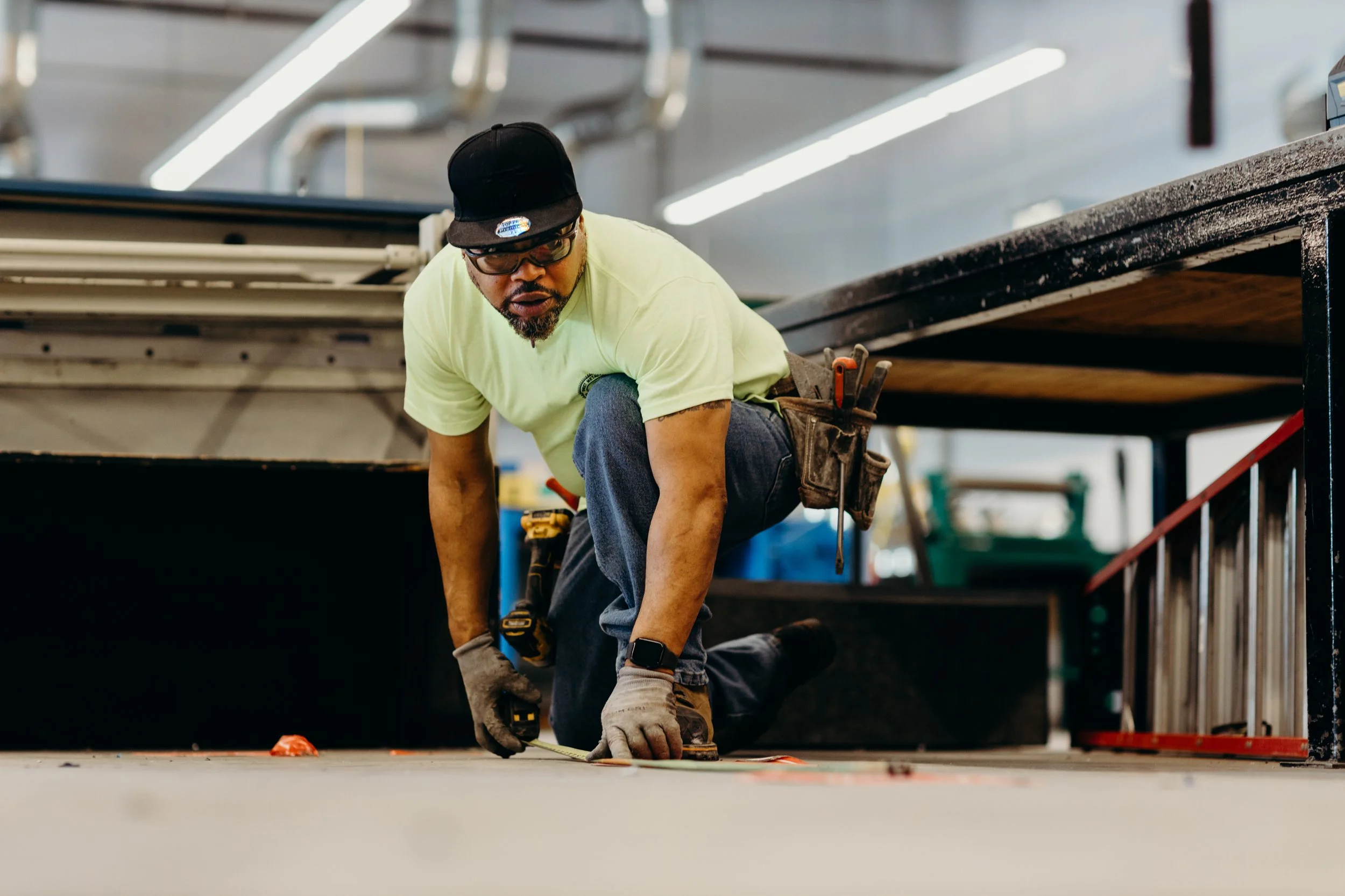 A man wearing a black cap, glasses, yellow T-shirt, jeans, and work gloves is kneeling on the floor using a measuring tape for construction or carpentry work in an industrial or workshop setting.