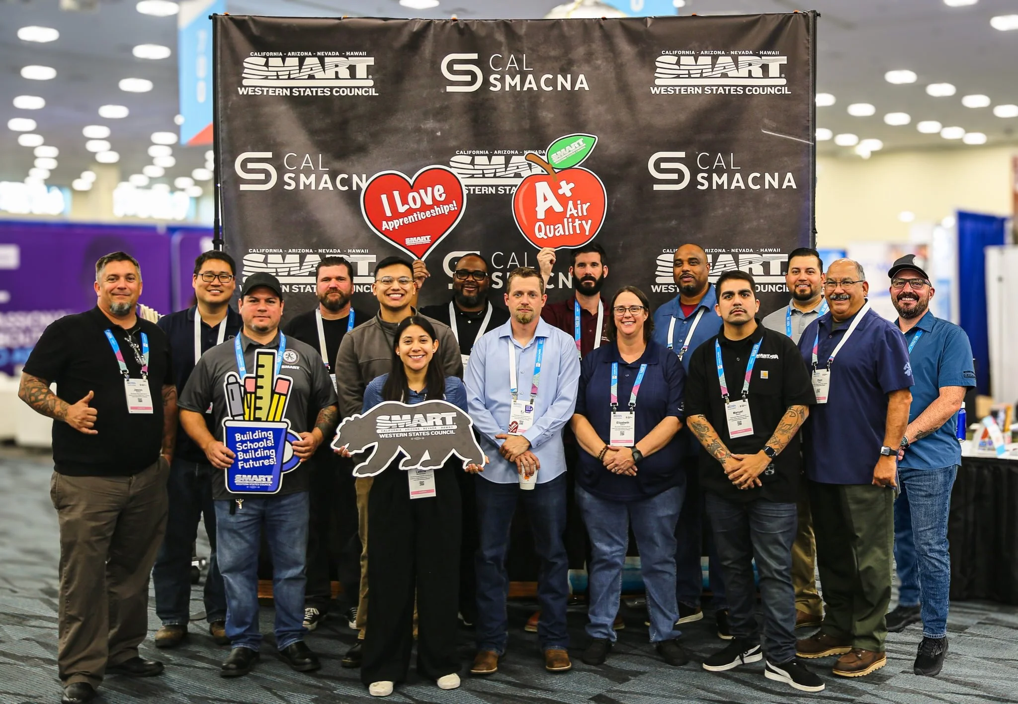 Group of people standing together at a conference, holding signs supporting apprenticeships and air quality, in front of a backdrop displaying logos for CAL SMART and Western States Council.