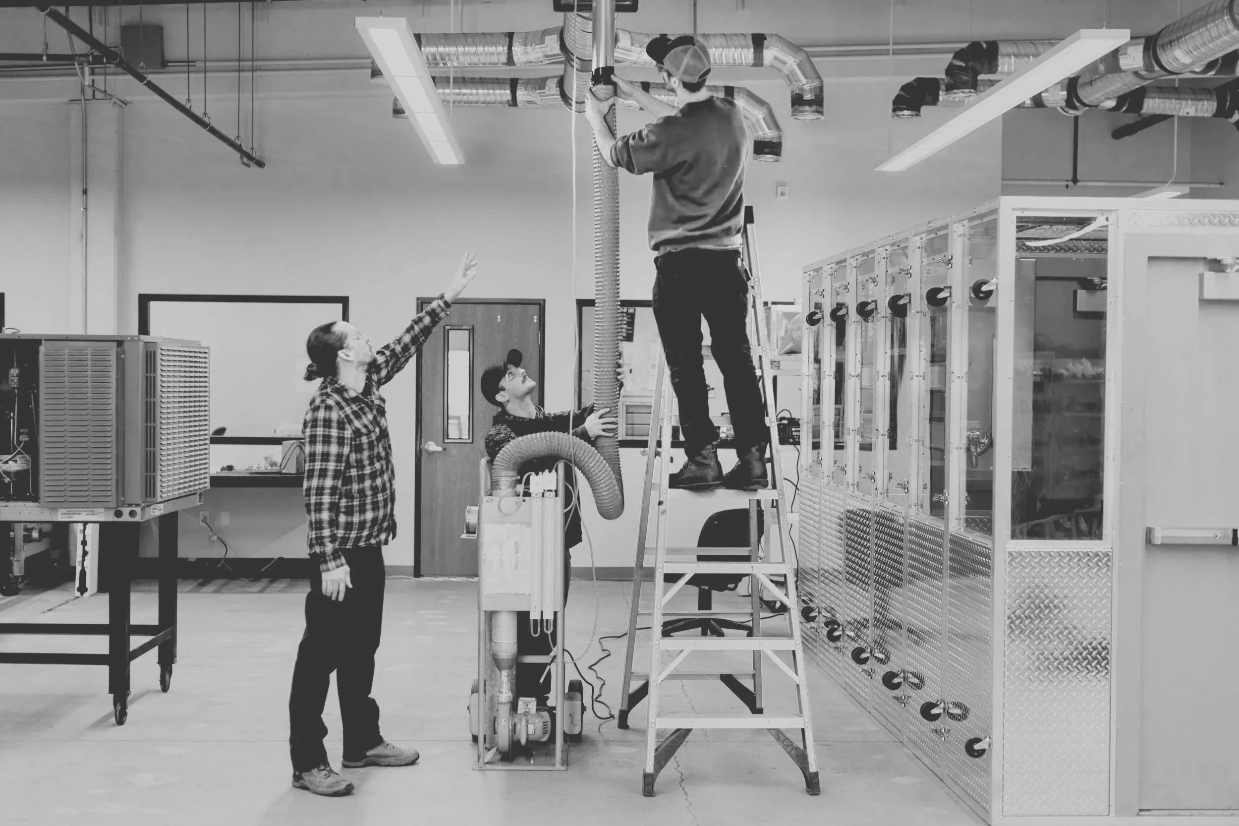 Three individuals working with HVAC equipment in an industrial or laboratory setting. One person is on a ladder adjusting ductwork, and the other two are assisting or supervising.