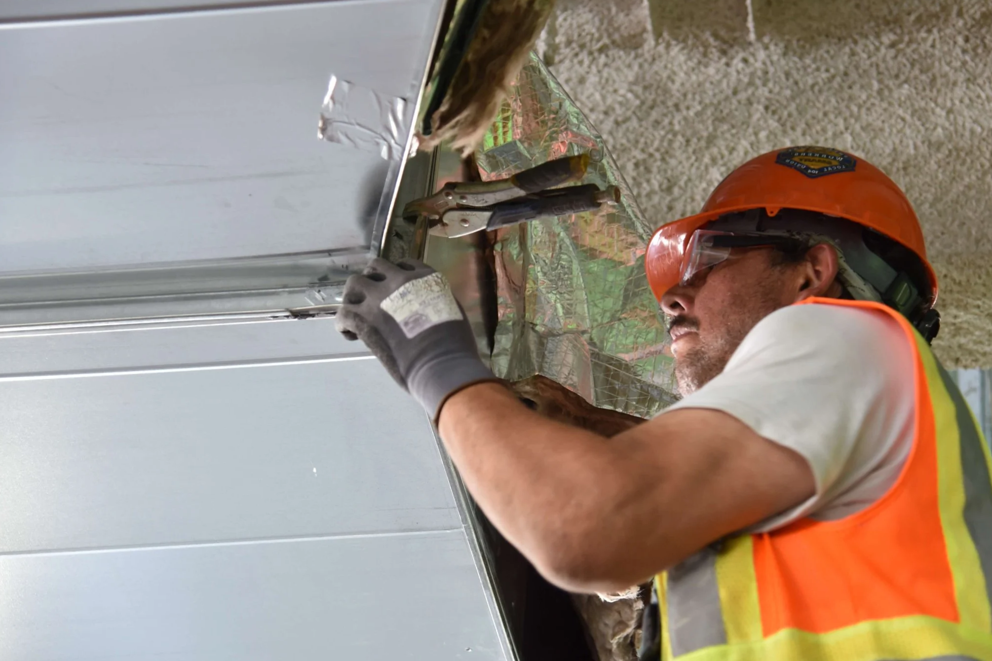 A construction worker with a beard, wearing an orange safety helmet, glasses, and a yellow safety vest, is installing or repairing a metal panel on a building facade.