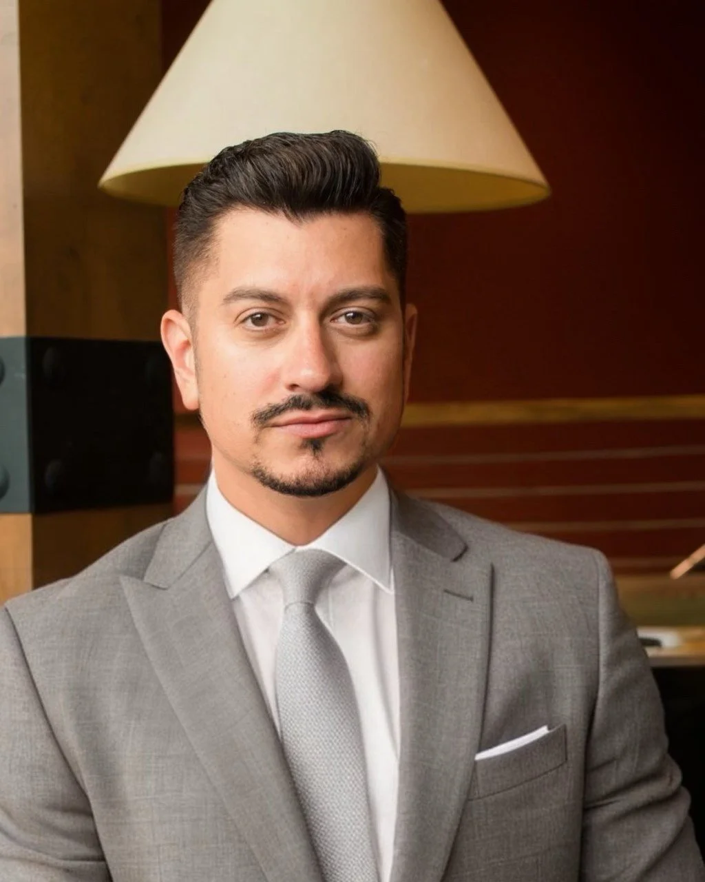 A portrait of Paul Thomas in a gray suit, white shirt, and light gray tie. He has dark hair, a goatee, and is sitting in front of a desk with a lamp in the background.