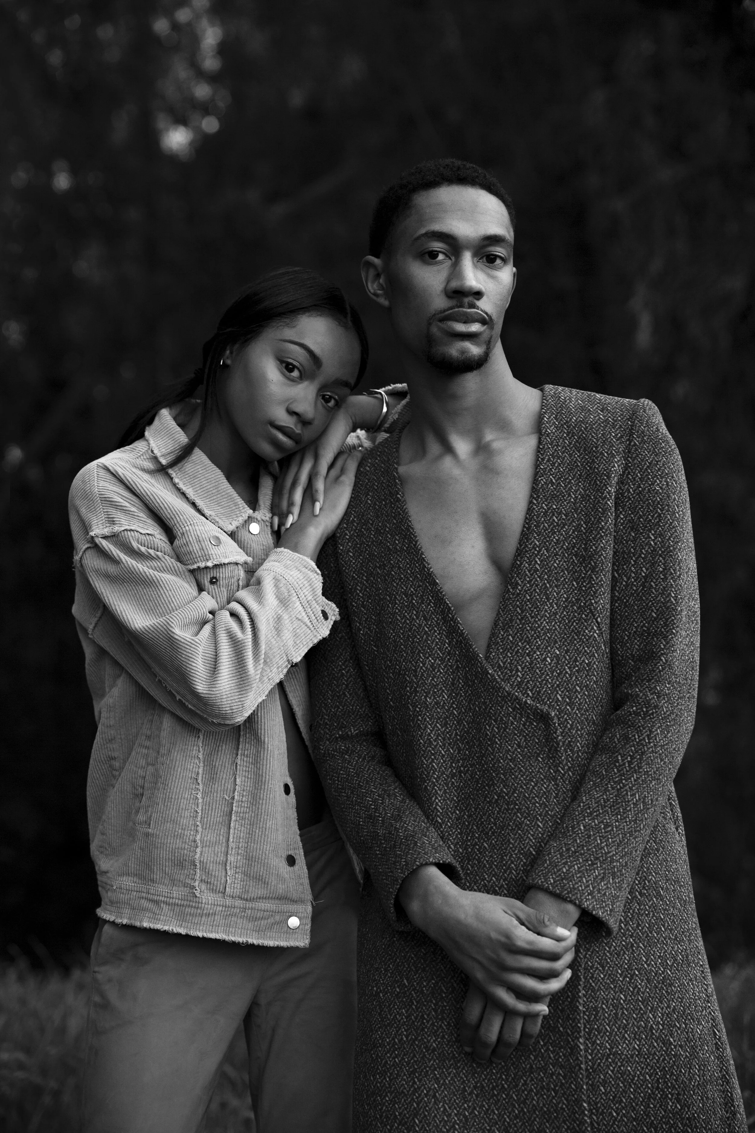 Black and white photo of a man and woman standing close together outdoors. The woman rests her head on the man's shoulder, and both are looking at the camera. The woman wears a light jacket and has her hand on the man's shoulder, while the man wears a textured blazer with his hands clasped in front of him. The background is blurred with dark foliage.