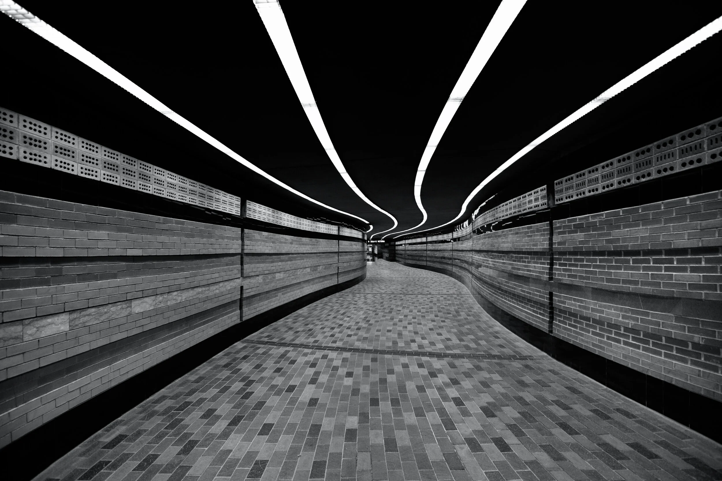 Curved underground pedestrian tunnel with brick walls, tiled floor, and modern ceiling lights.