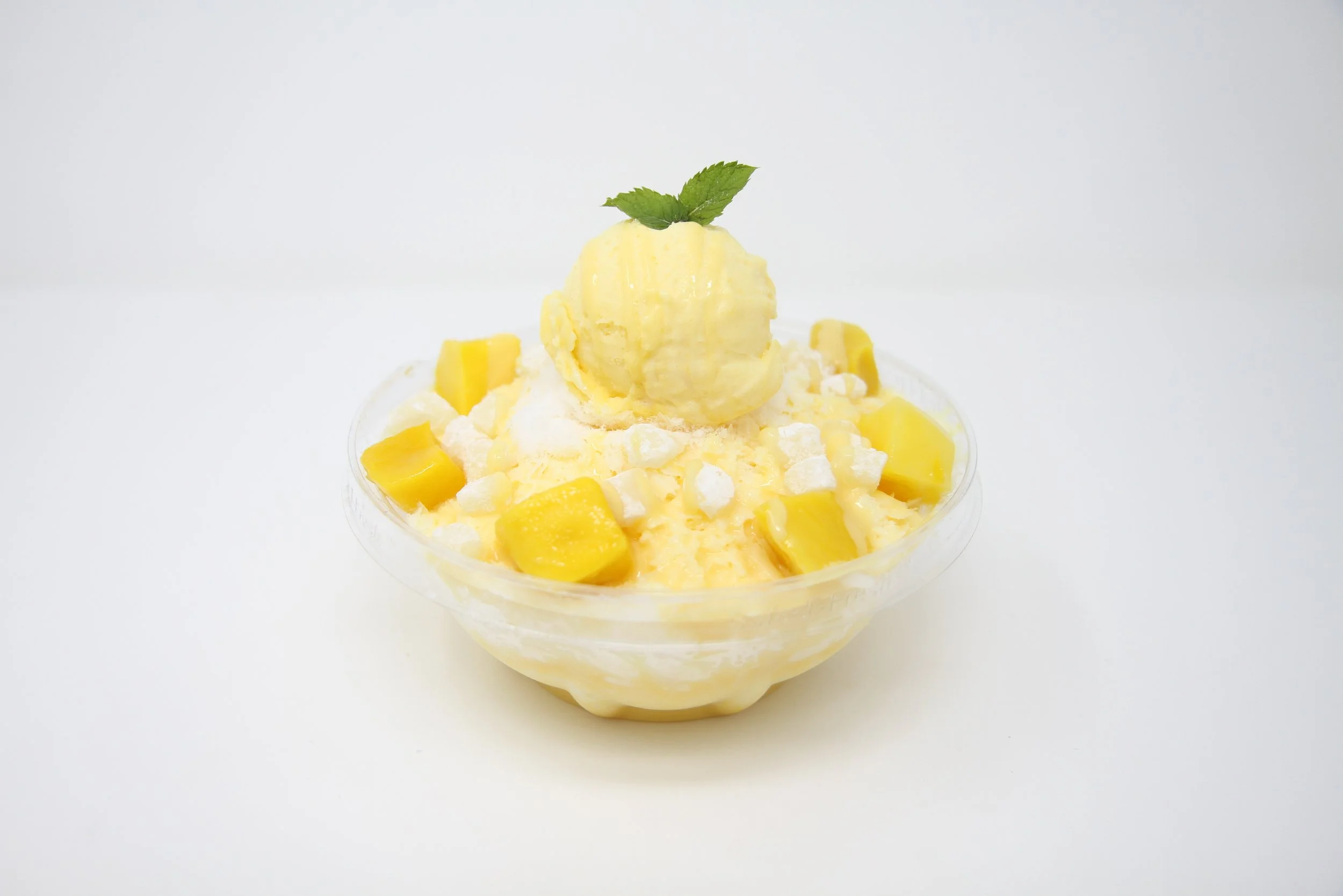 Mango Milk Snow in clear plastic bowl, layered shaved milk ice, mango condense milk, mango fruit pieces, mochi, and mango gelato.