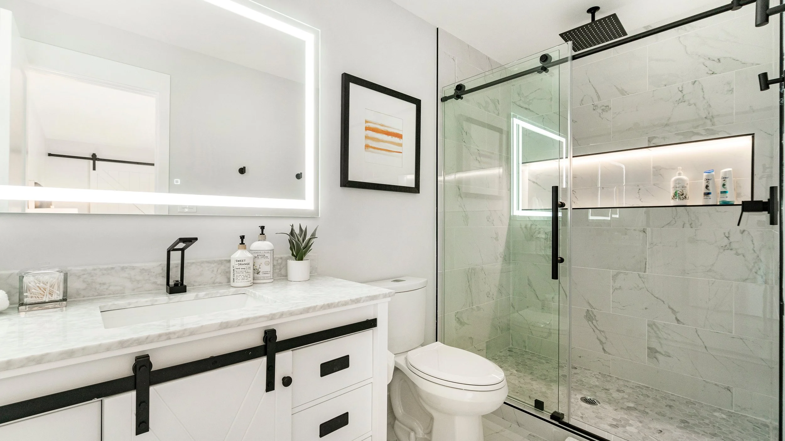 Modern bathroom with a large illuminated mirror, a framed artwork, white marble countertop with soap dispensers and a potted plant, a white toilet, and a glass walk-in shower with marble tiles and built-in shelf holding toiletries.