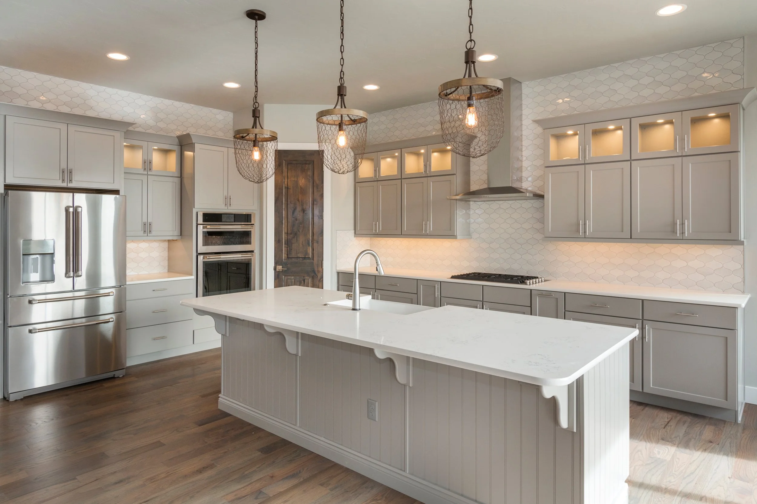 Modern kitchen with gray cabinets, white marble island, stainless steel refrigerator, double oven, stovetop, wood flooring, pendant lights, and neutral-toned backsplash.