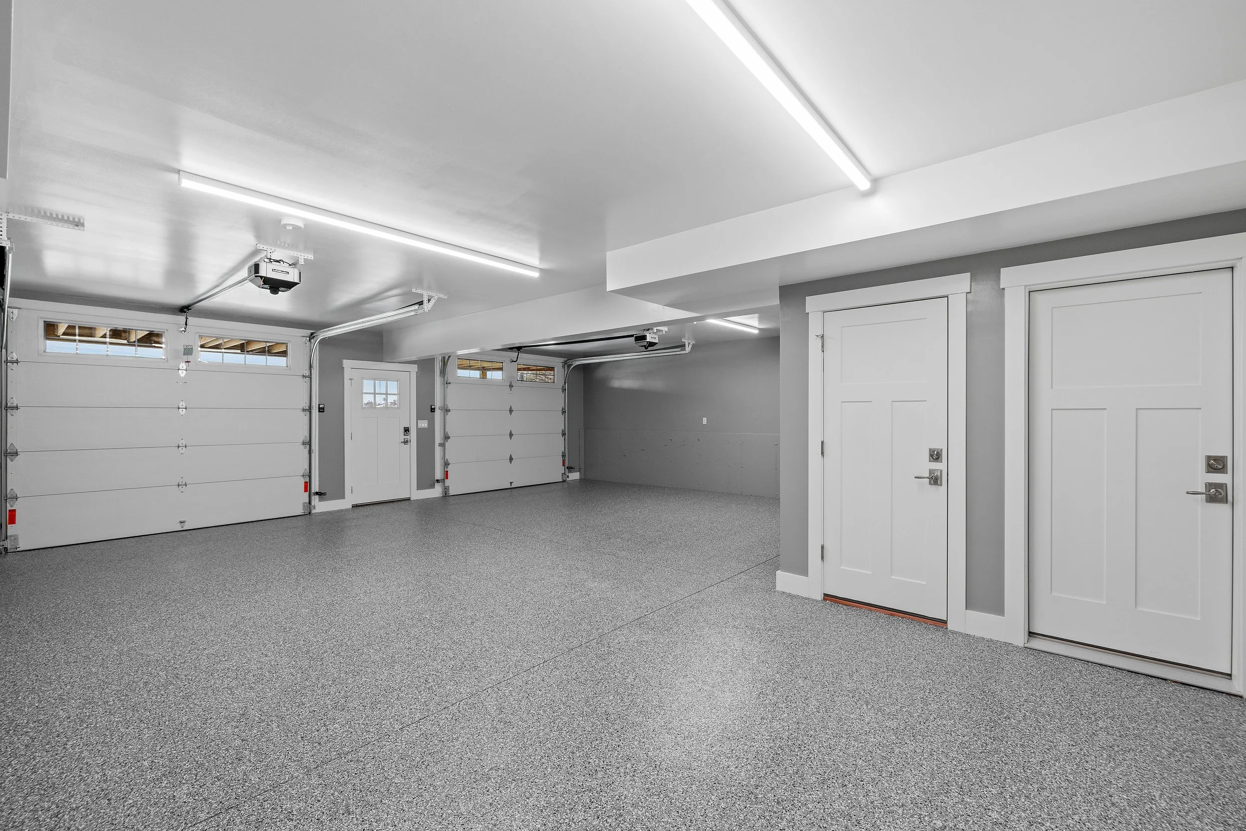 Empty garage with two white sectional garage doors, a side door, gray walls, and bright LED lighting.