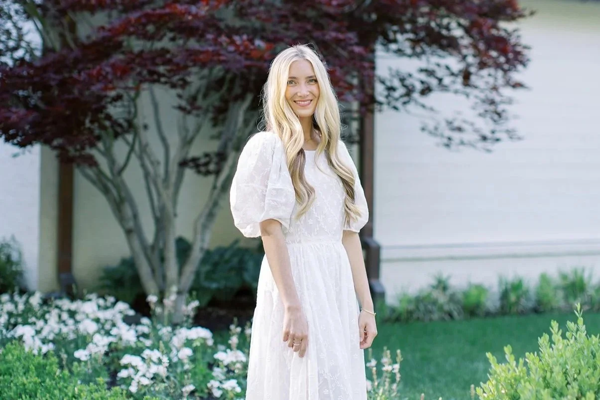 A young woman with long blonde hair, smiling, wearing a white dress with puff sleeves standing outdoors in front of a tree with dark red leaves and a house with white siding.