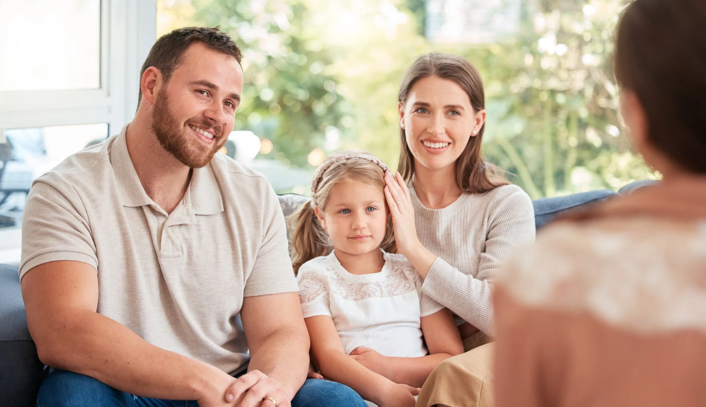 A family of four sitting on a couch, engaging in a conversation with a woman who is facing them. The family includes a man, a woman, and two young girls, with a bright, sunlit outdoor scene visible through the window behind them.