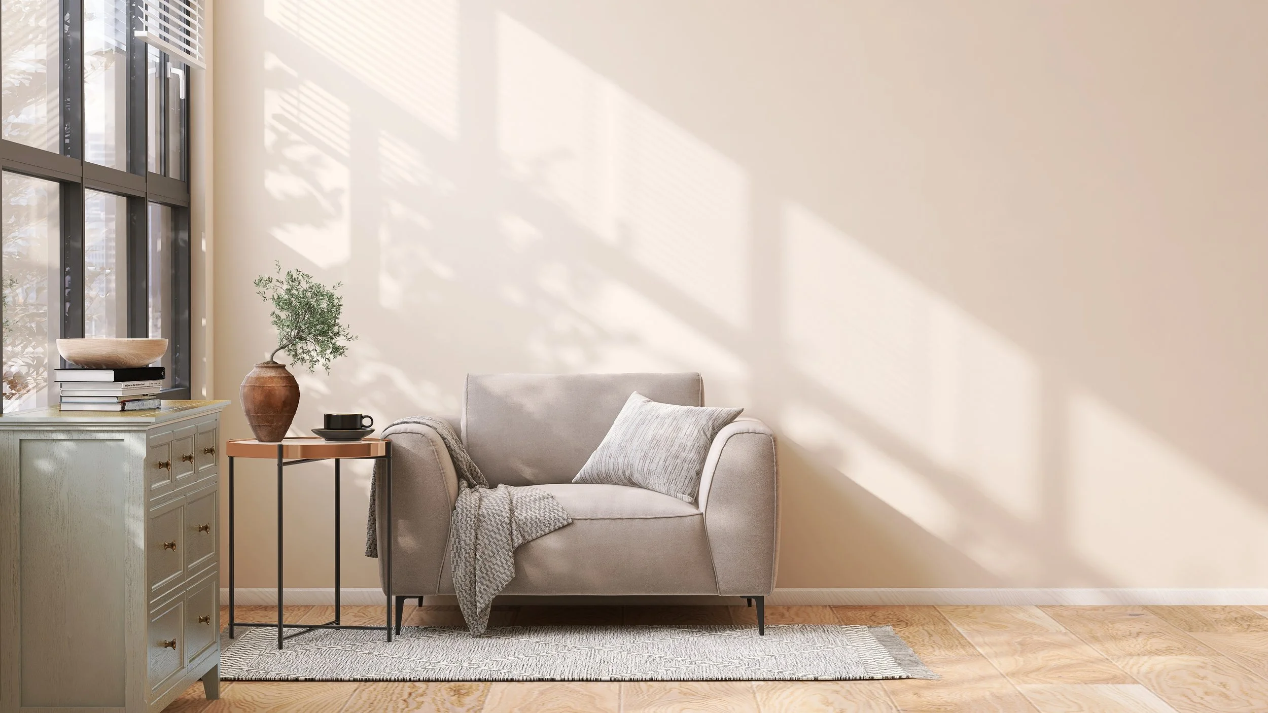 Sunlit living room with cream-colored sofa, grey pillows, beige wall, and light wooden flooring. A white sideboard has books and a wood bowl; a small side table holds a black cup and saucer, and a tall potted plant. Shadows cast by window blinds.