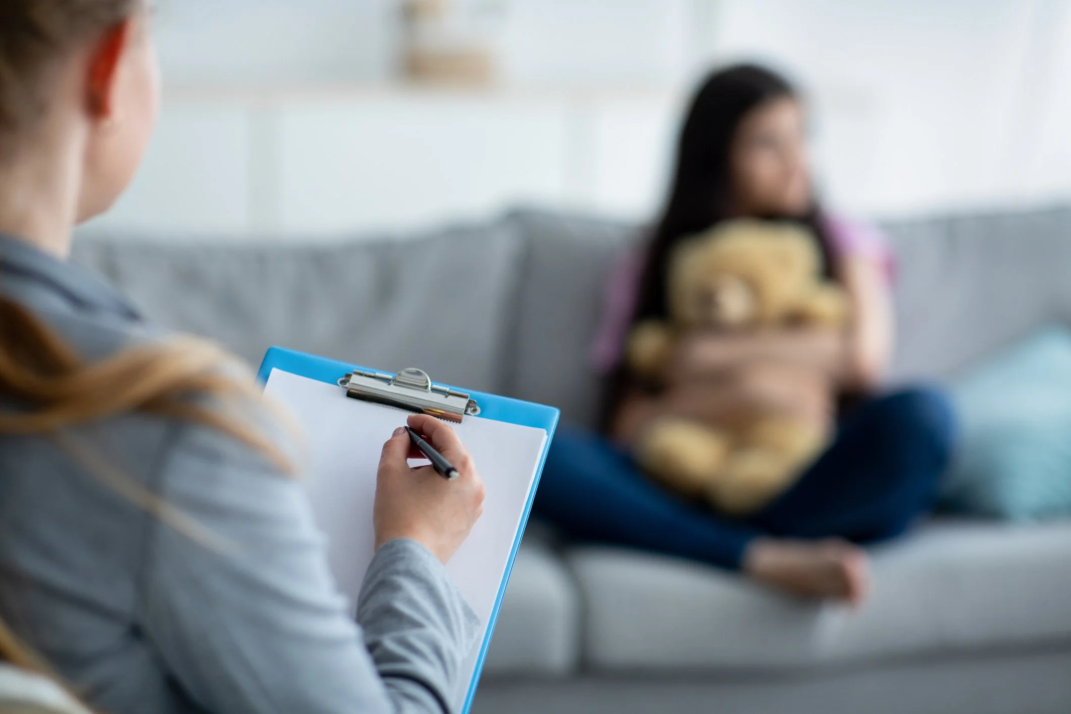 A woman with long hair writing on a notepad with a pen while sitting in a waiting room, with a blurred woman in the background holding a teddy bear.