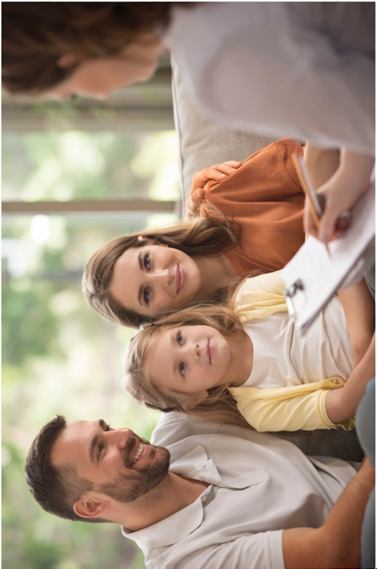 A family of three sitting on a couch, smiling and looking at a laptop. The family includes a man with a beard, a woman with long brown hair, and a girl with blonde hair, in a bright room with large windows and green outdoor scenery in the background.