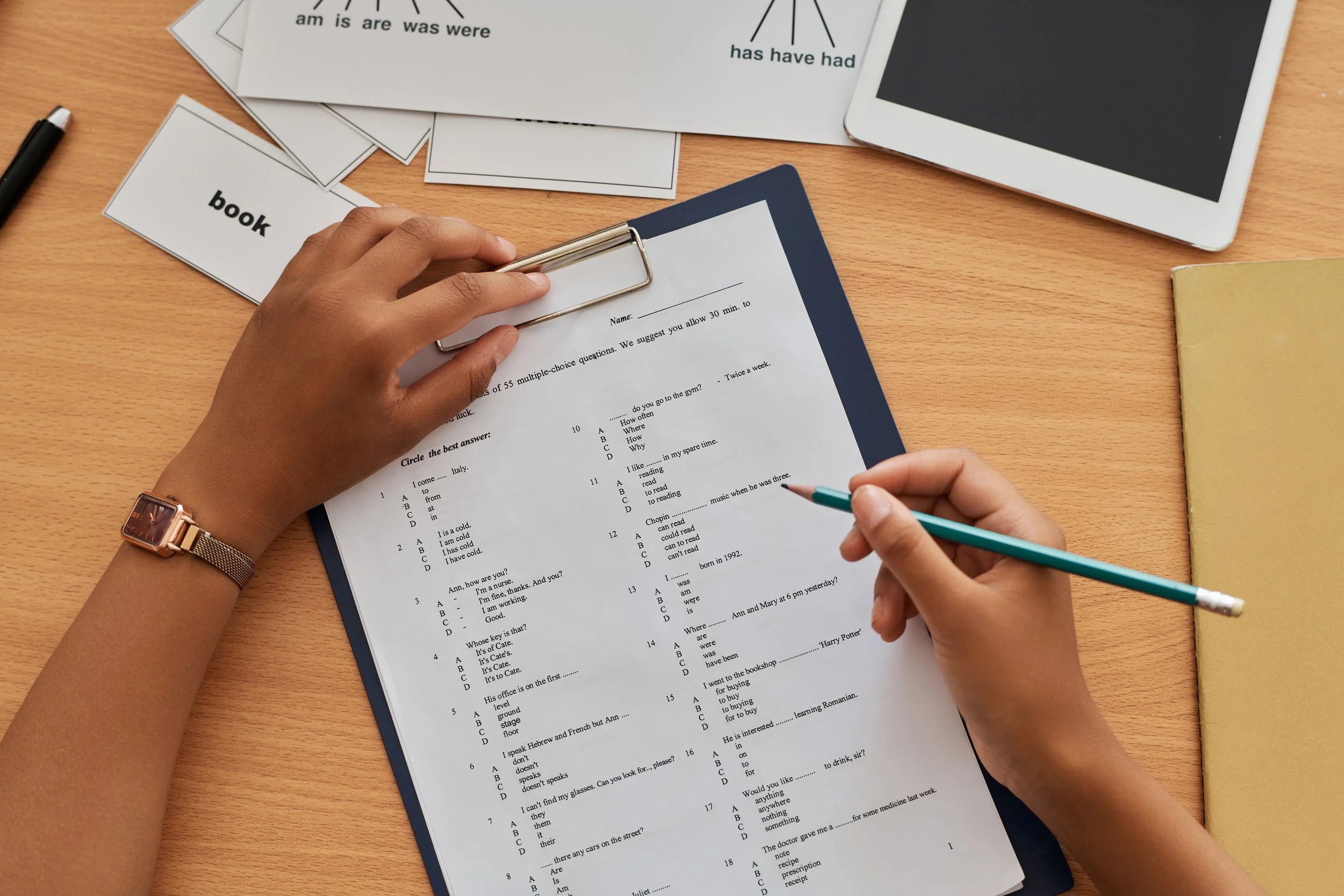 A person's hands holding a clipboard with a multiple-choice test and a pen, on a wooden table with scattered flashcards and a tablet.