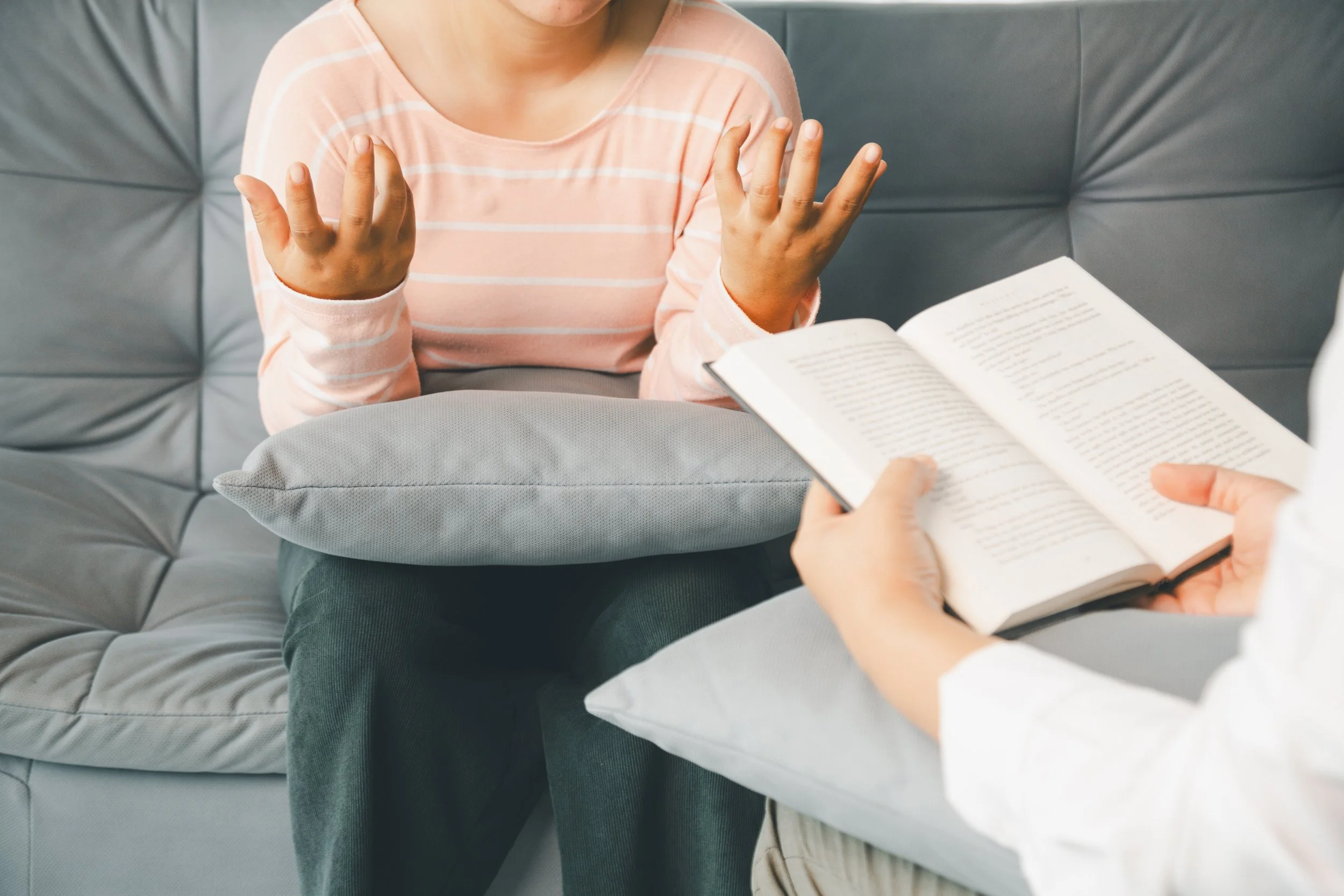 Person gesturing with hands while sitting on a gray cushioned bench, with an open book in front of them.