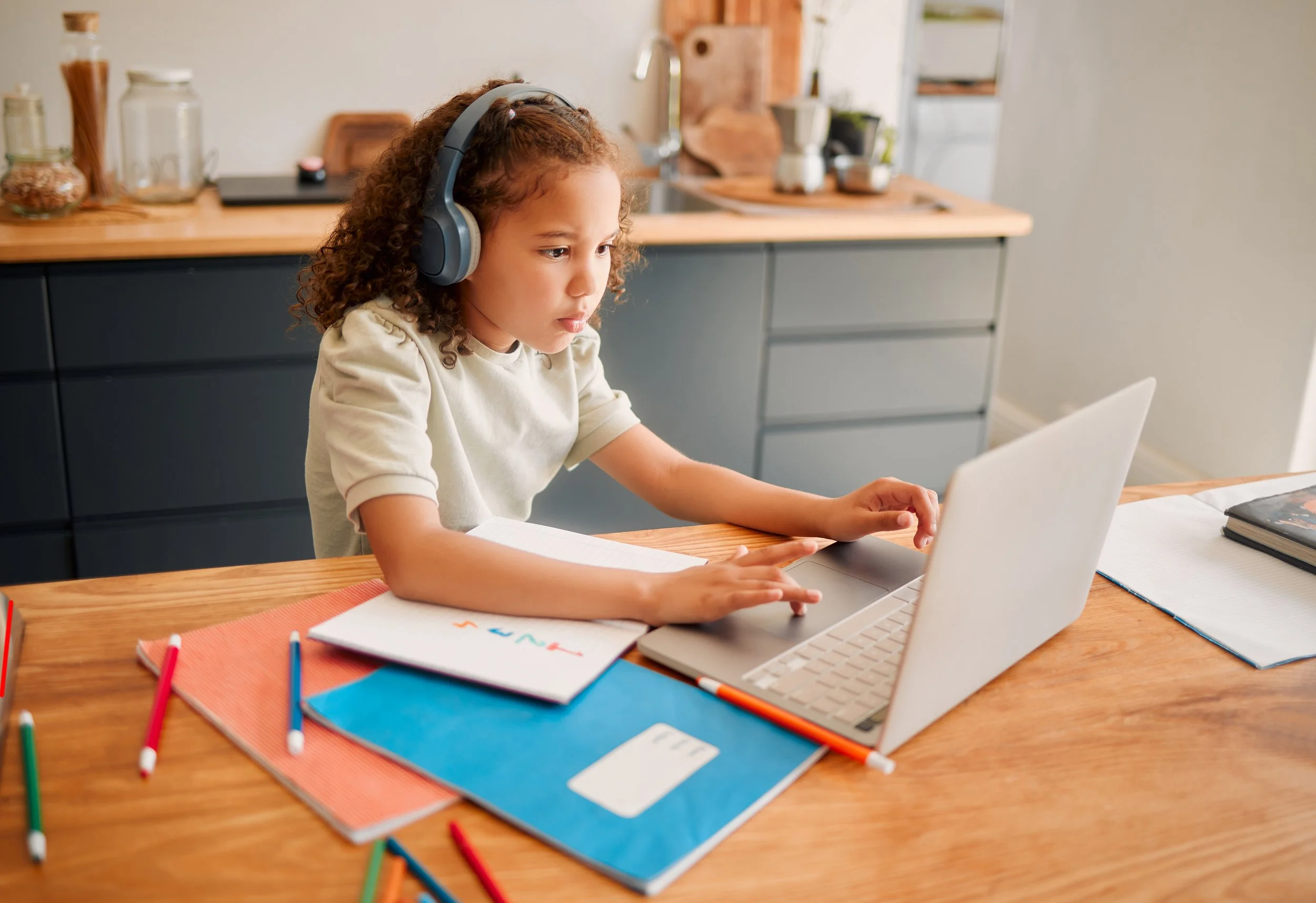 A young girl with curly hair wearing headphones, working on a laptop at a wooden table with notebooks and colorful pens, in a kitchen with jars and a sink in the background.