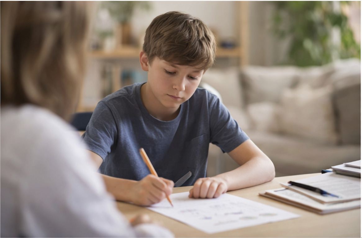A young boy doing homework or coloring at a wooden table with notebooks and papers, sitting in a cozy living room with beige pillows and a plant in the background.