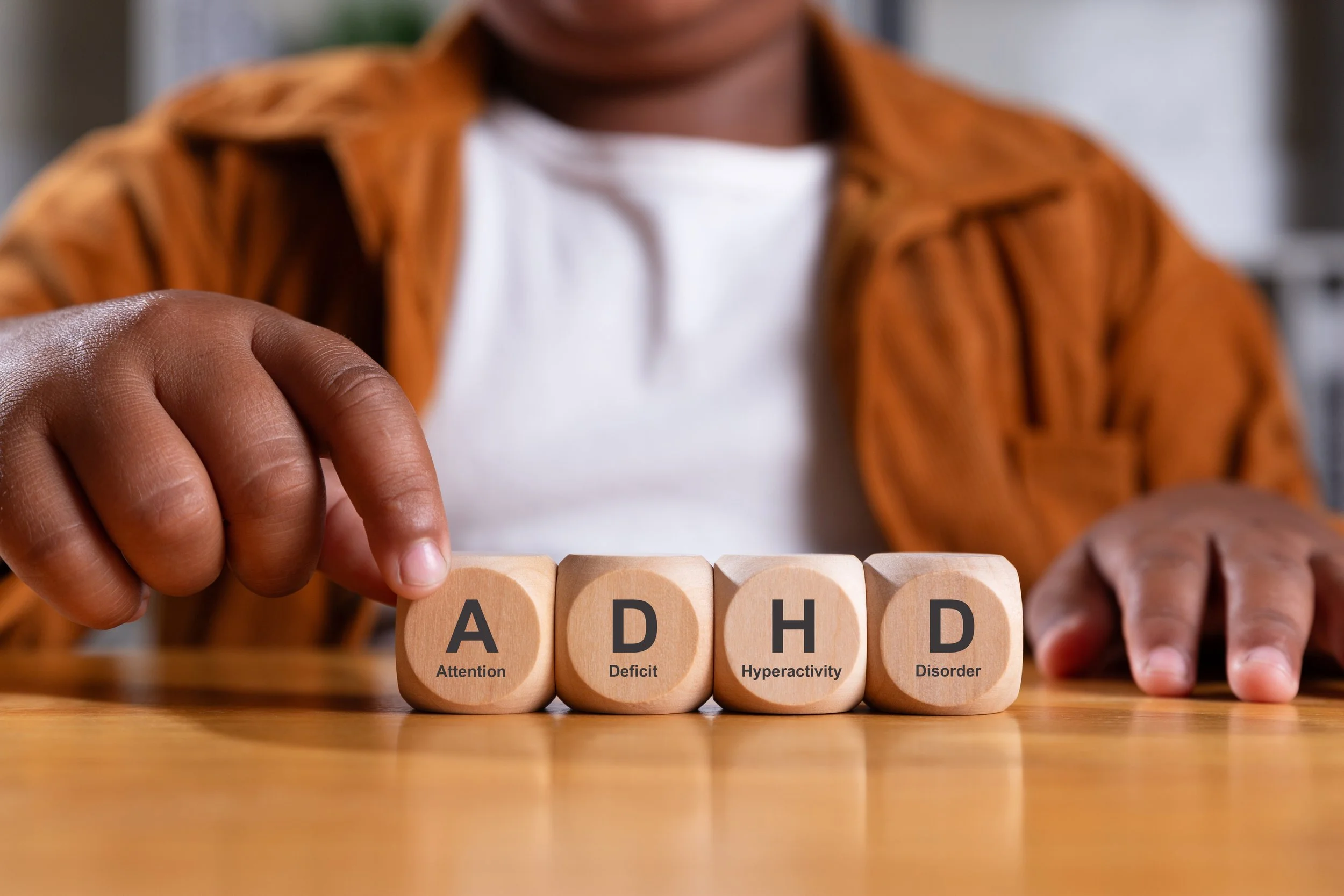 Close-up view of a person's hand with dark skin touchingly holding a wooden block with an 'A' on it, labeled 'Attention' with other blocks labeled 'Deficit', 'Hyperactivity', and 'Disorder' behind, in focus with a blurred background.