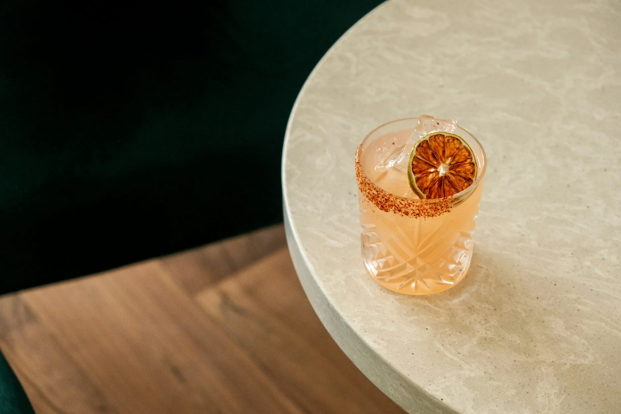 A glass with a pink drink garnished with a slice of dried lime on ice, placed on a round, light-colored stone table.