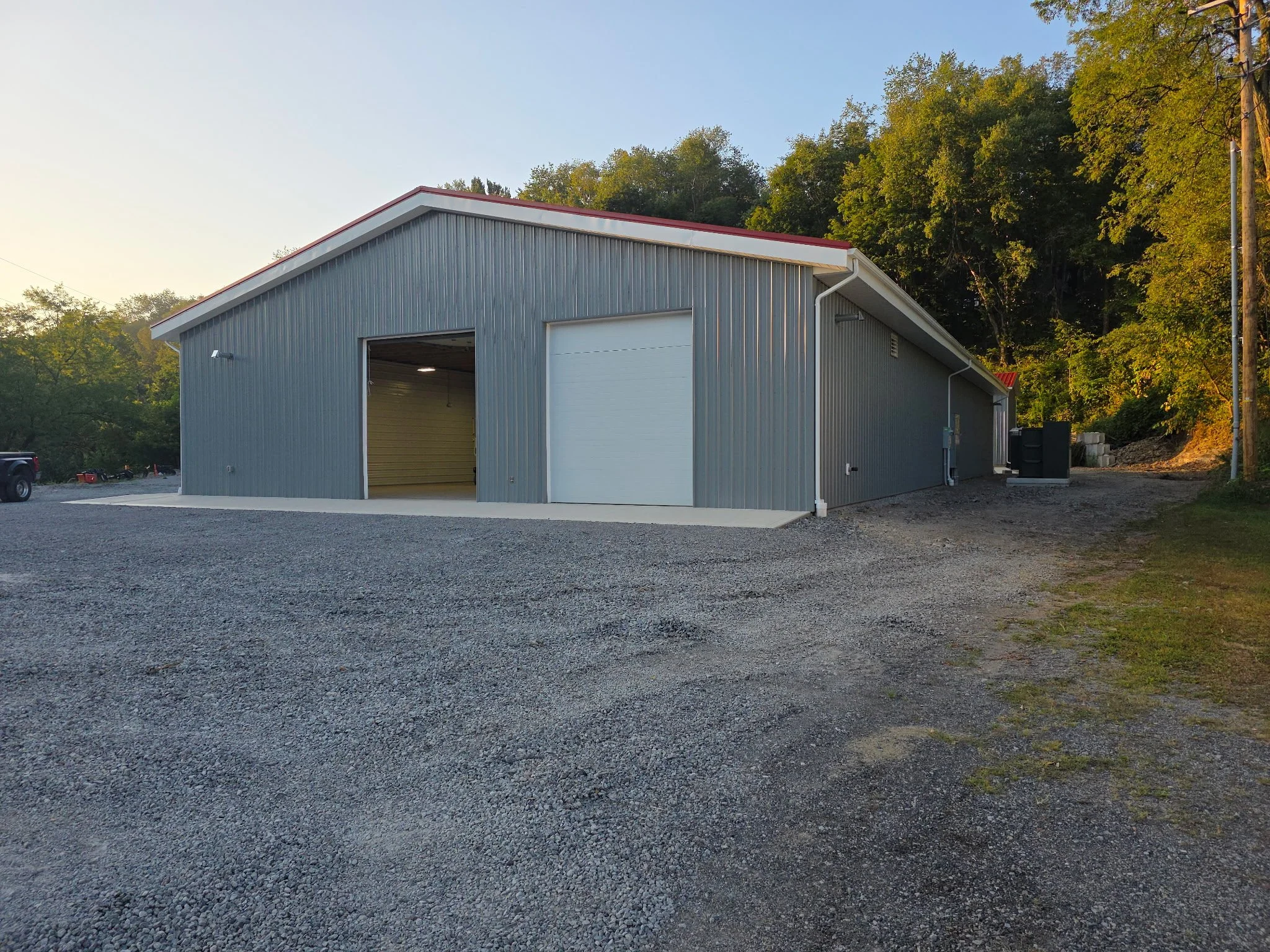 A large metal building with a peaked roof, gray siding, and a white roll-up garage door, situated on a gravel lot with trees in the background.