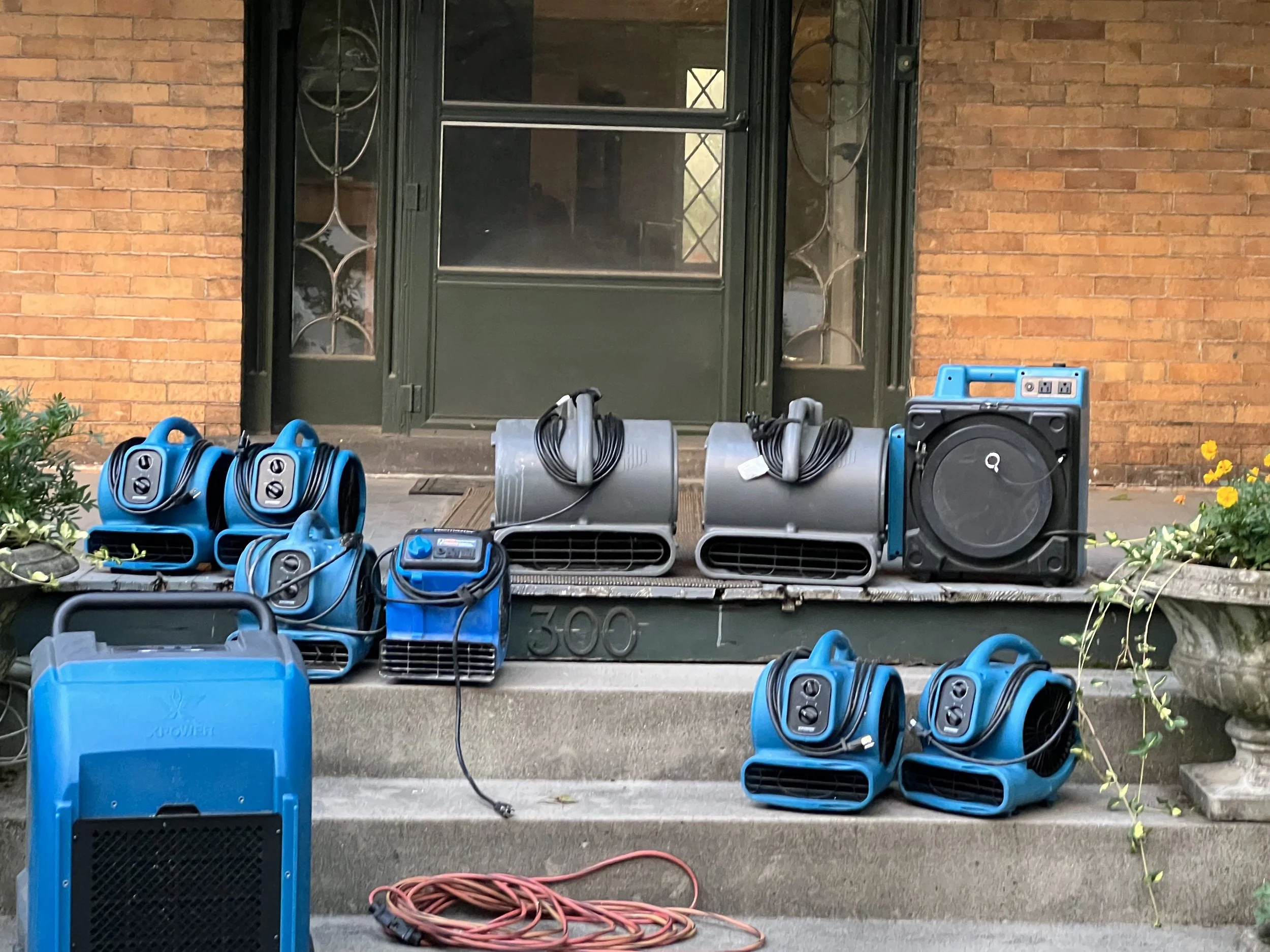Collection of portable air coolers and fans in blue and gray placed on front stairs of house with brick wall and green front door.