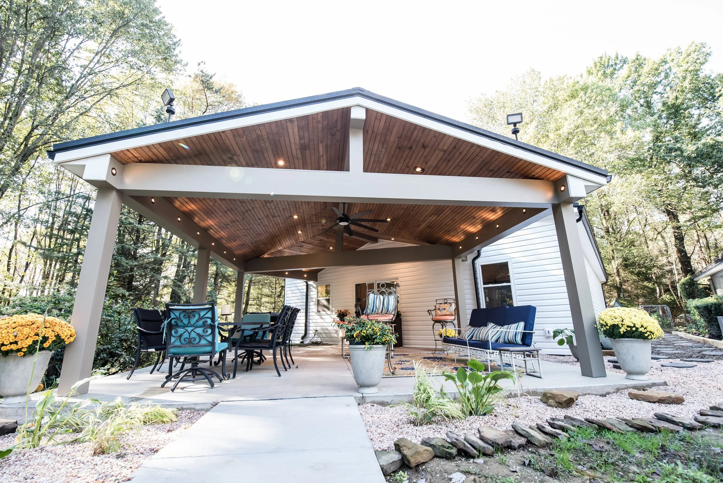 Backyard patio area with outdoor furniture, flower pots, and a wooden ceiling, surrounded by trees.