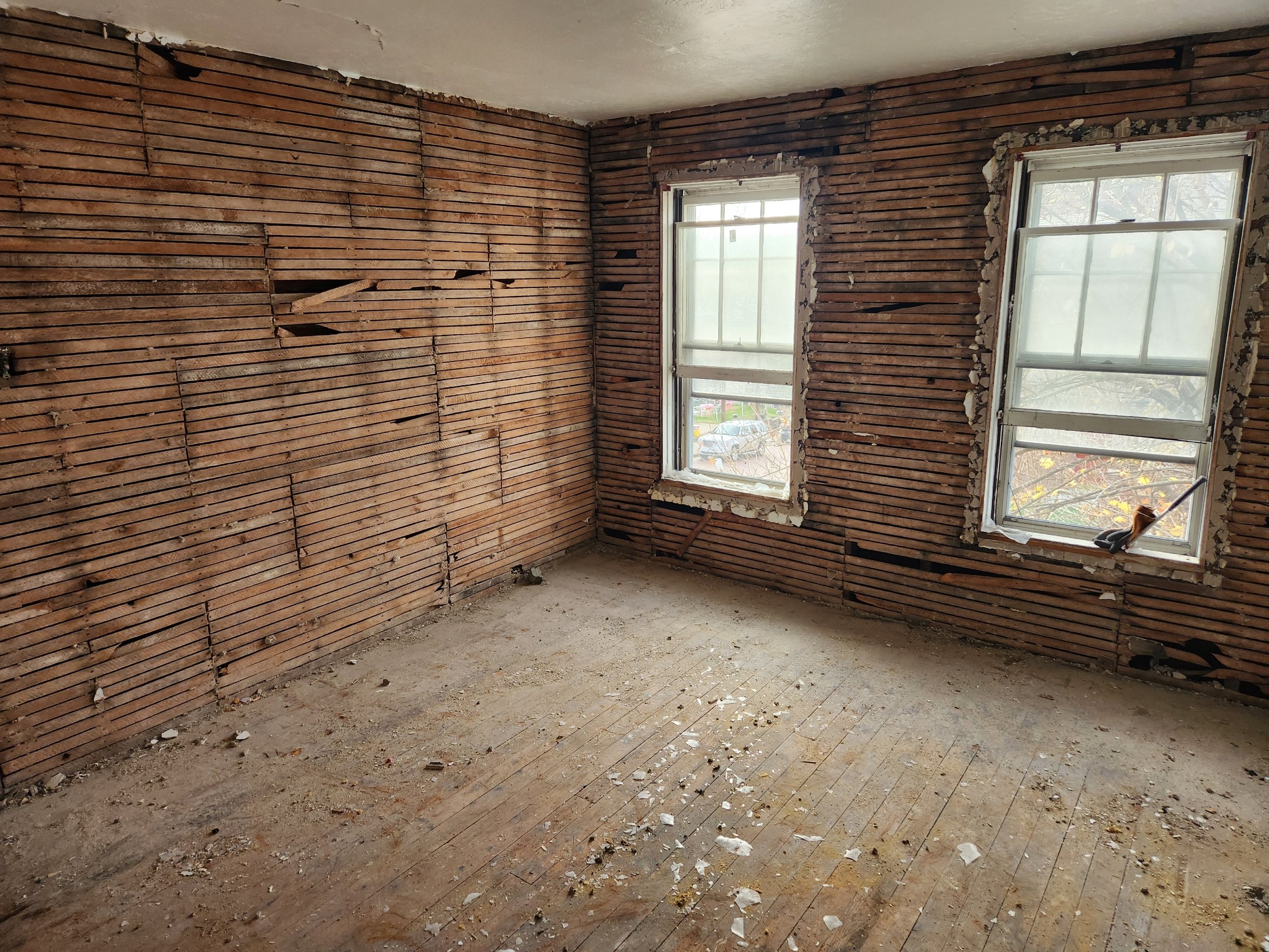 Empty room with exposed brick walls, two windows with peeling paint, dirt and debris on the floor, and construction tools in the window