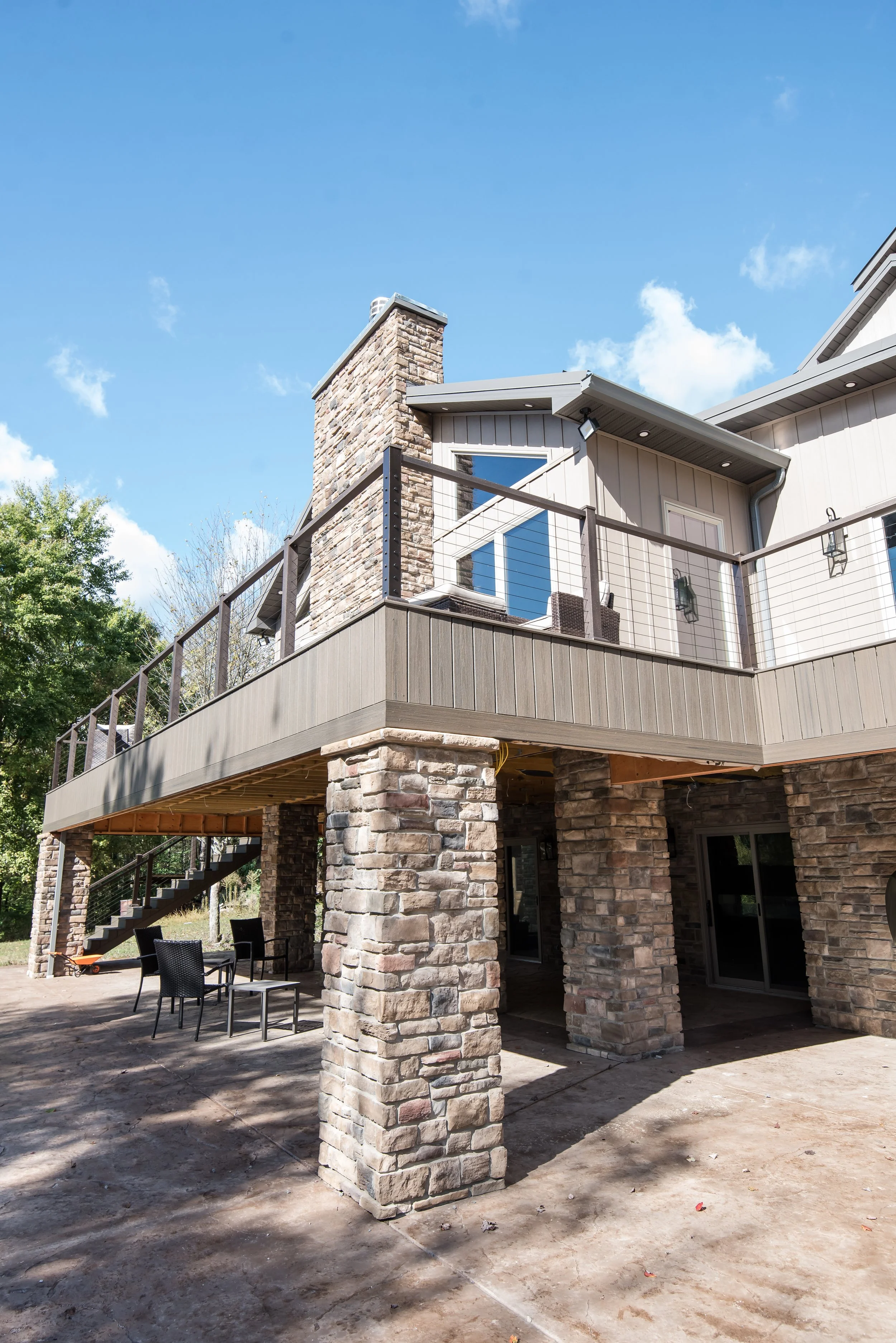 Exterior view of a two-story house with a stone and wood facade, featuring a balcony with a black railing, outdoor seating area with chairs, and a staircase leading to the upper level, under a clear blue sky.