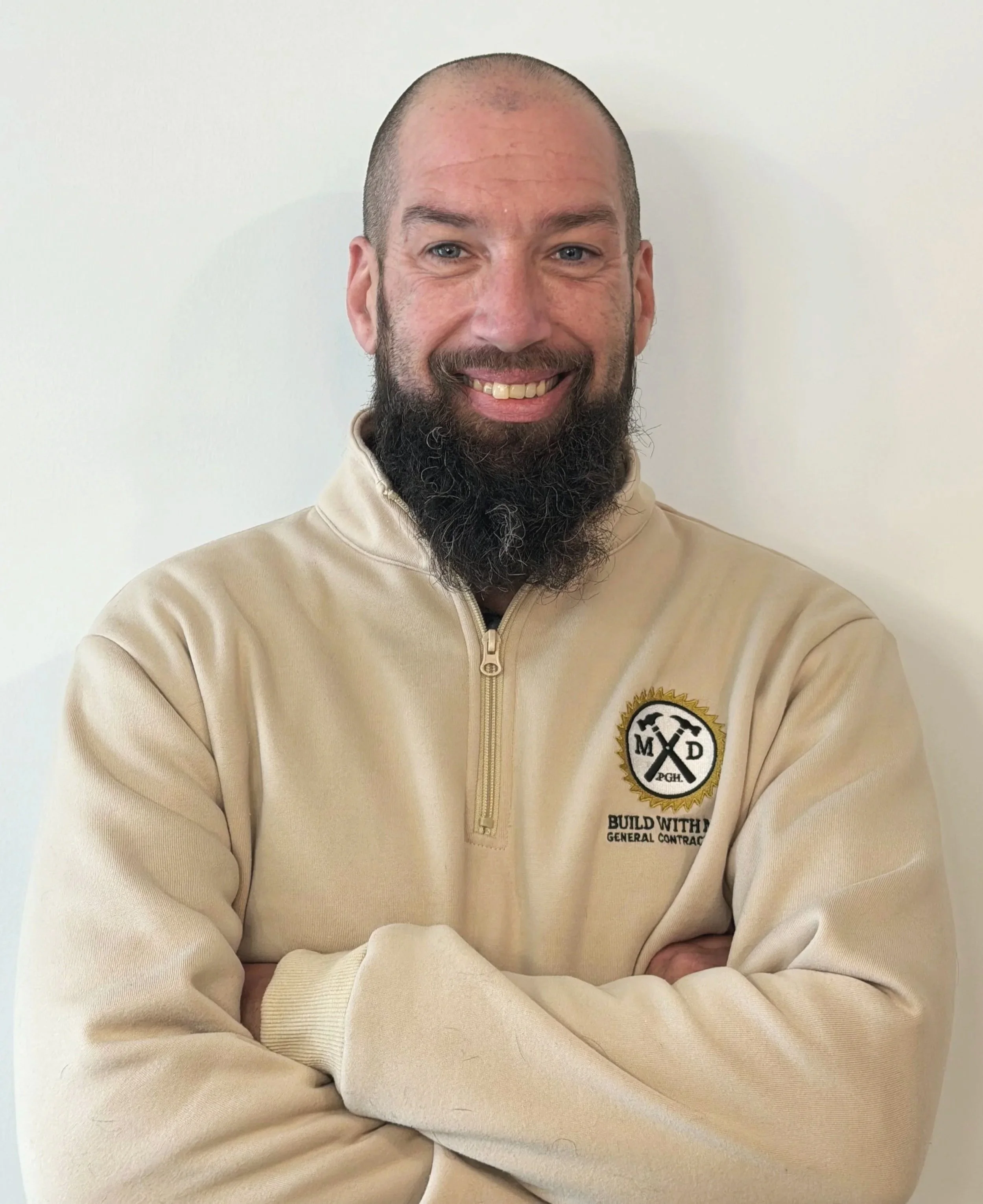 A man with a beard and a smile, standing with arms crossed, wearing a beige sweatshirt with a logo on it, against a plain background.